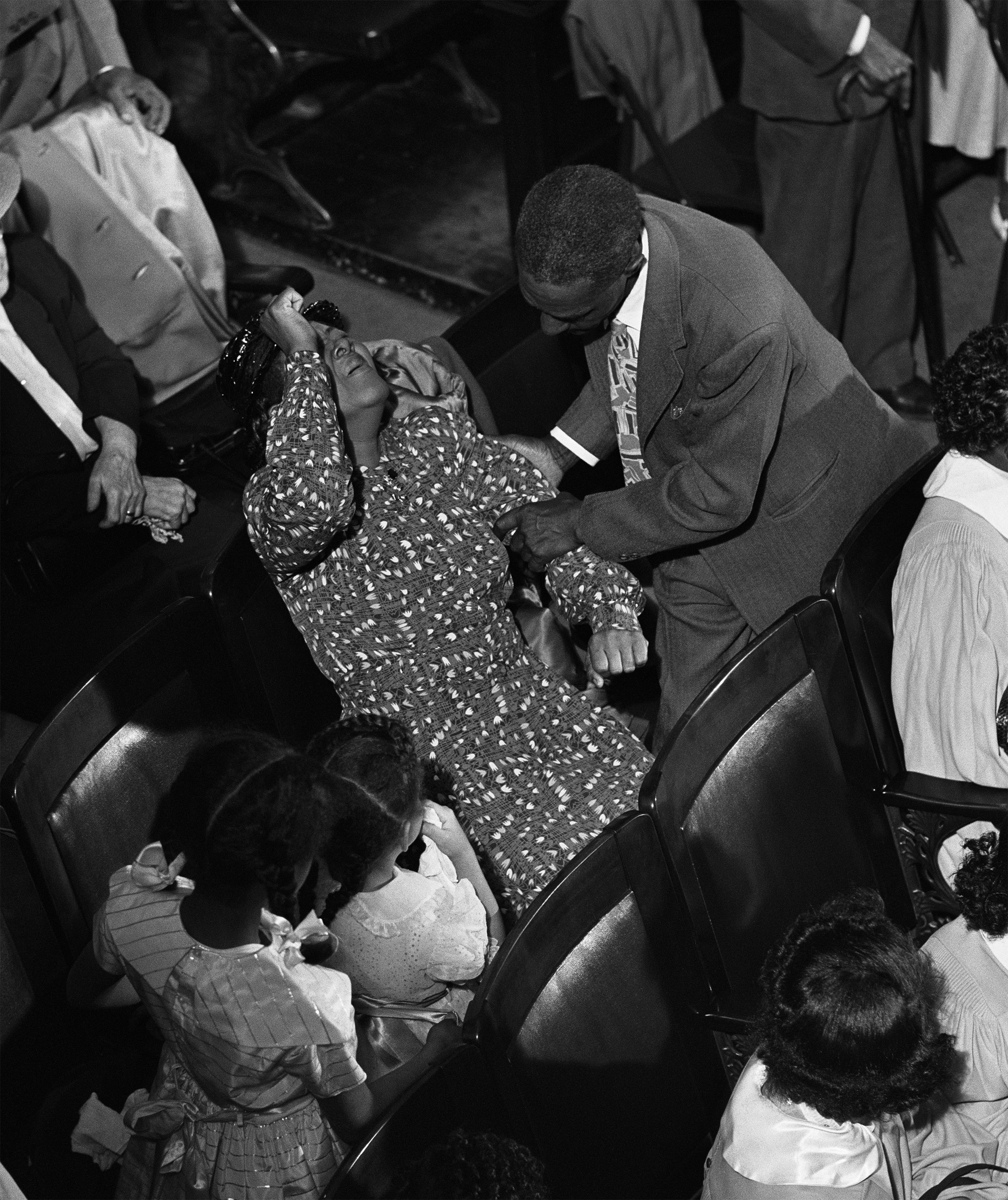 A woman in a patterned dress reclines in a wooden theater-style seat with one hand raised to her forehead, supported by a man in a suit leaning over her. Several people, including young girls in dresses with ribbons and women in white blouses, sit or stand nearby, watching or attending. The scene suggests an emotional or spiritual response, likely taking place in a church or auditorium during a religious service.