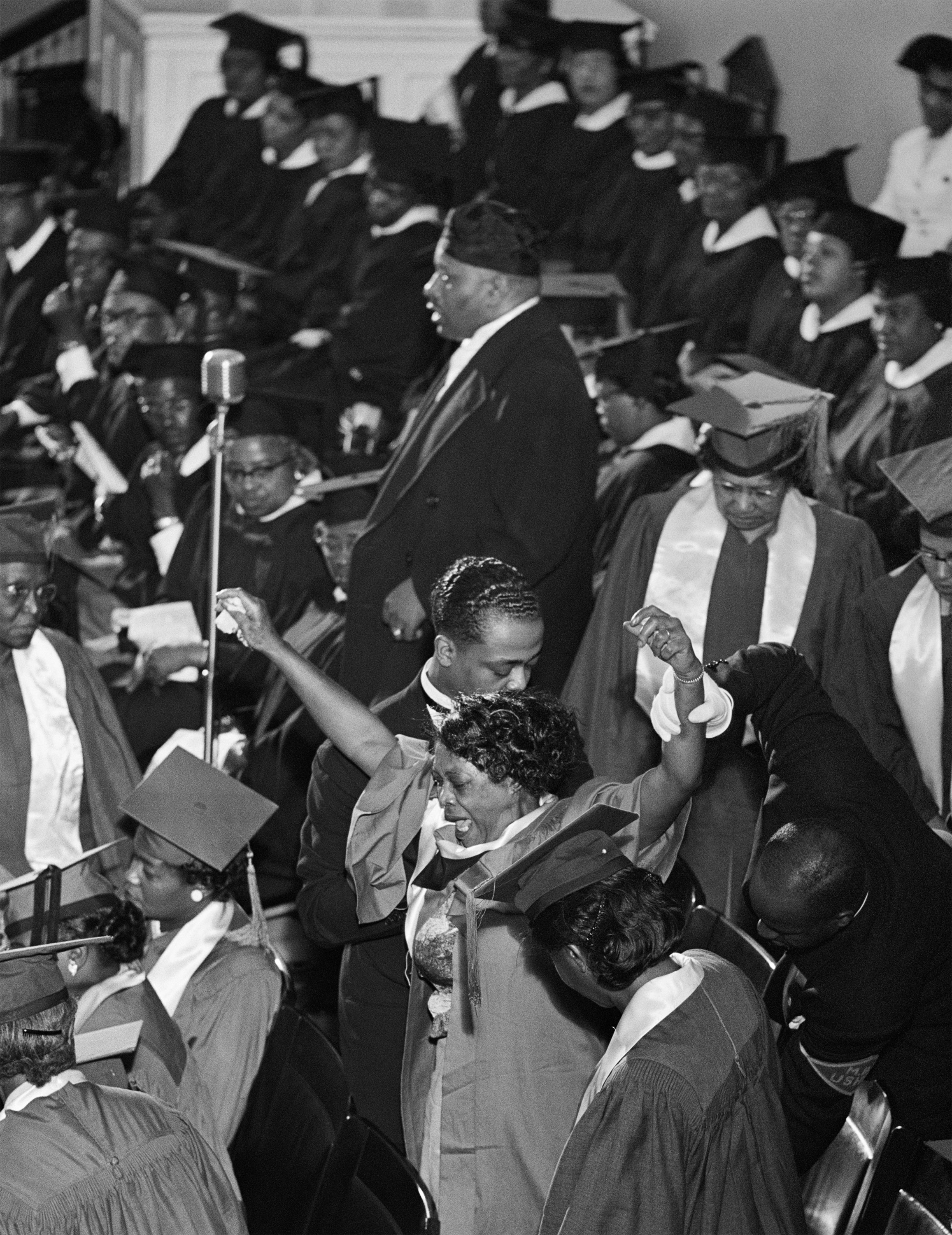 Congregants in caps and gowns fill a church, singing and reacting to a religious or ceremonial moment. At center, a woman in graduation robes raises both arms, her face expressive with joy or praise. She is supported by two men at either side, one helping her stand. Rows of seated women in robes surround her, some looking toward her while others read or sing. A large choir fills the background, also in robes and mortarboards, with a man standing in front of them beside a tall microphone.