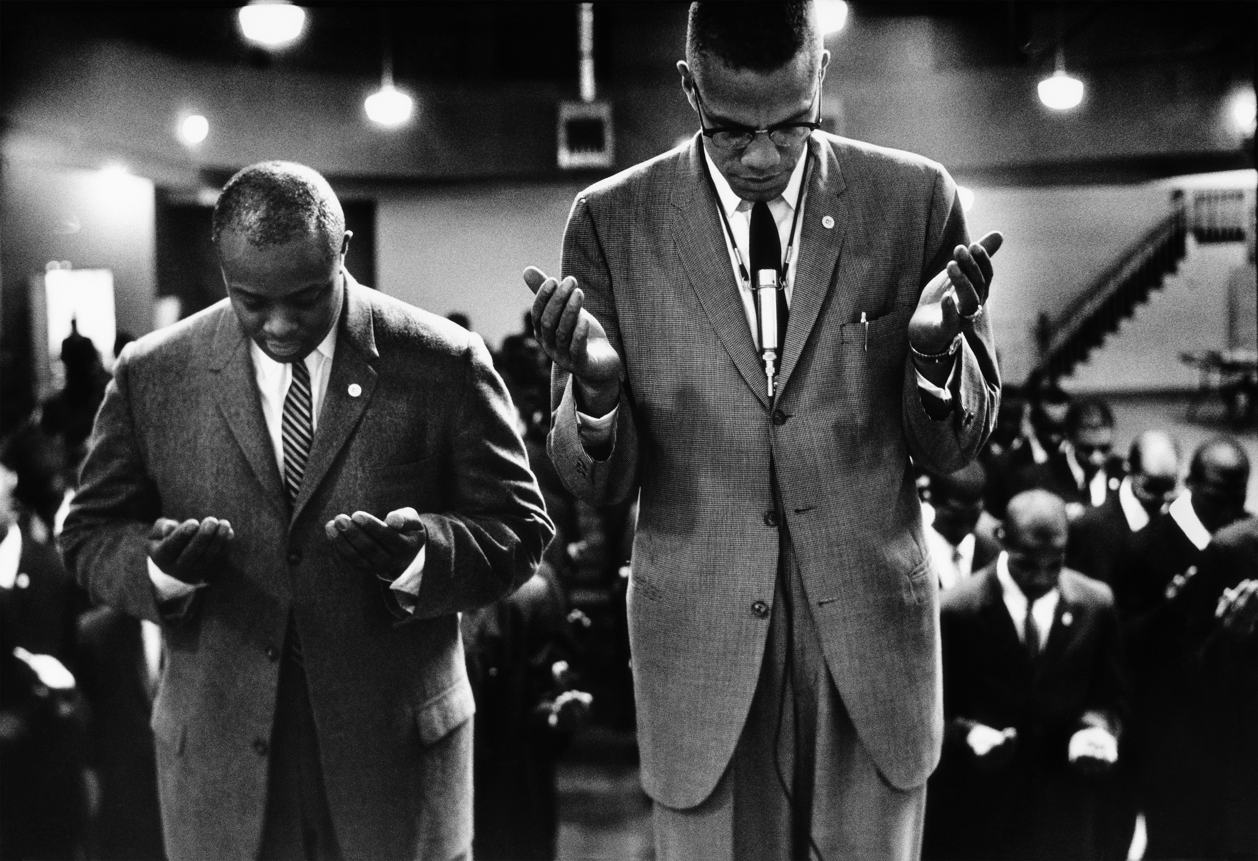 Two men in suits stand at the front of a large group of people, heads bowed and hands raised in prayer. The man on the right wears glasses, a pinstripe suit, and a visible tie clip with a pen in his breast pocket; the man on the left wears a striped tie and a pin on his lapel. Rows of similarly dressed men in suits stand behind them, also in prayer. The setting appears to be an indoor auditorium, with overhead lights and a staircase in the background.