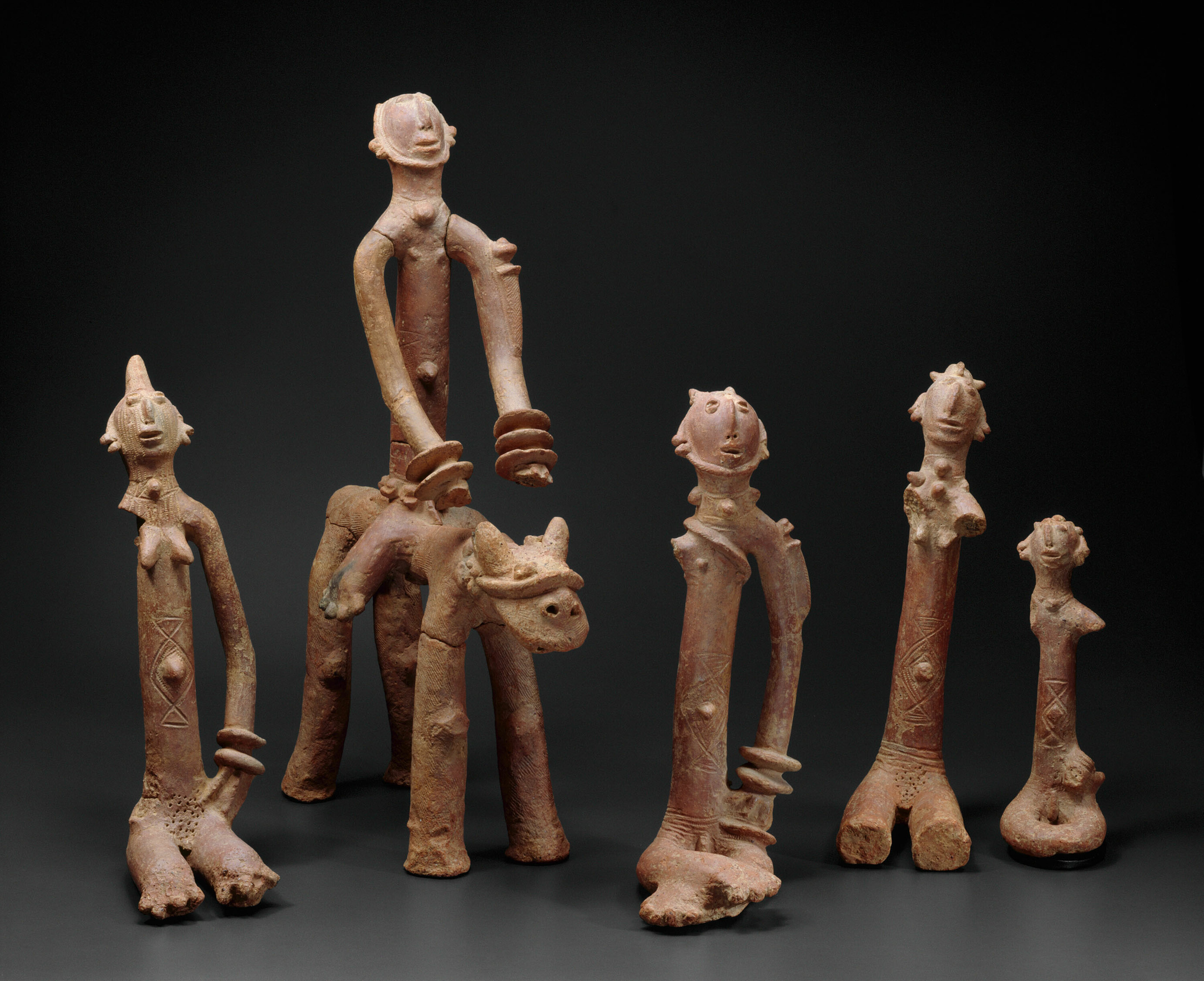 Five terracotta sculptures of abstract figures adorned with traditional hairstyles and possible scarification marks. Four are standing, while one rides a horse and has a knife strapped to his left arm.