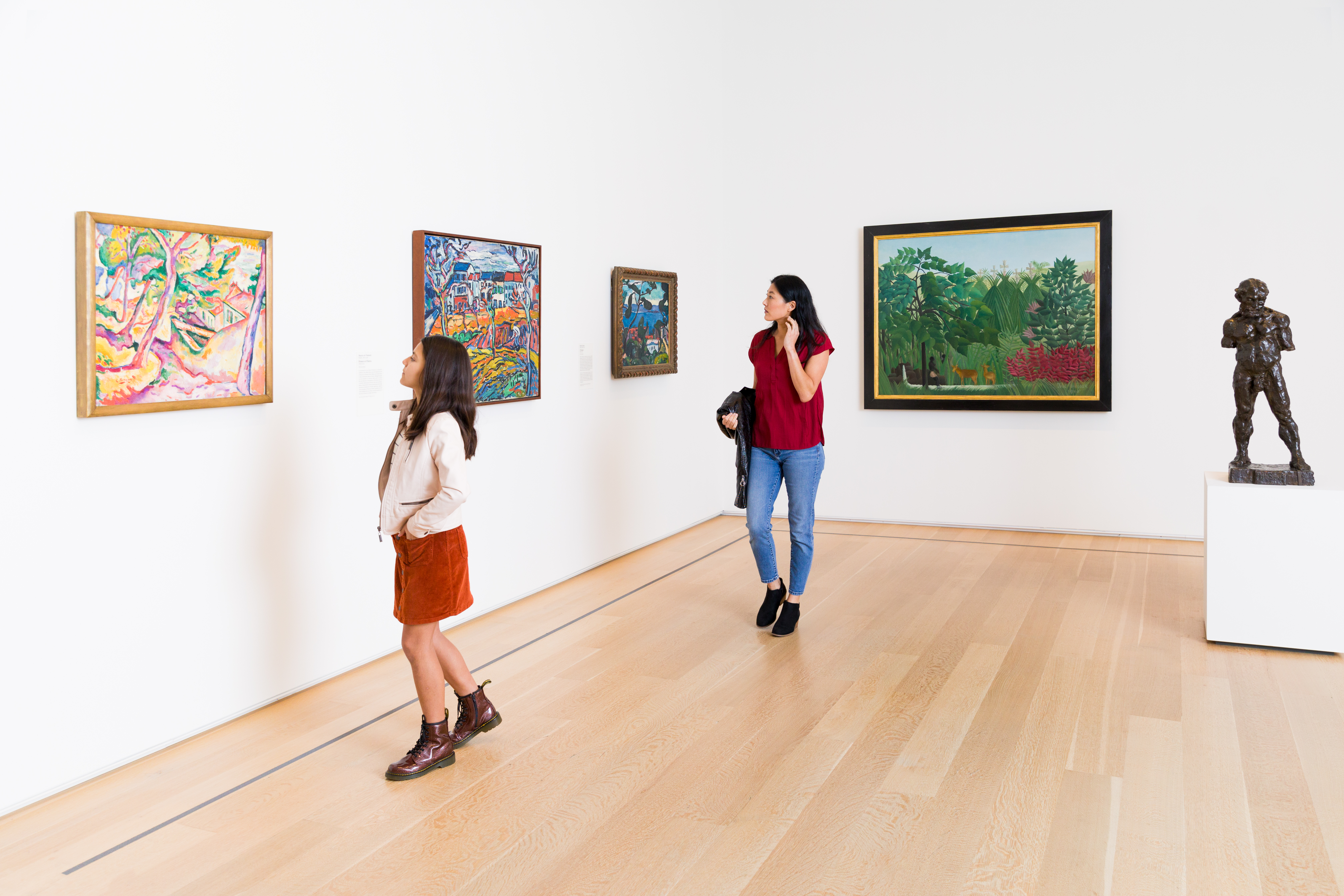 Photo of a teenage girl with brown hair and a young woman with black hair viewing brightly colored Fauvist paintings in a white art gallery.