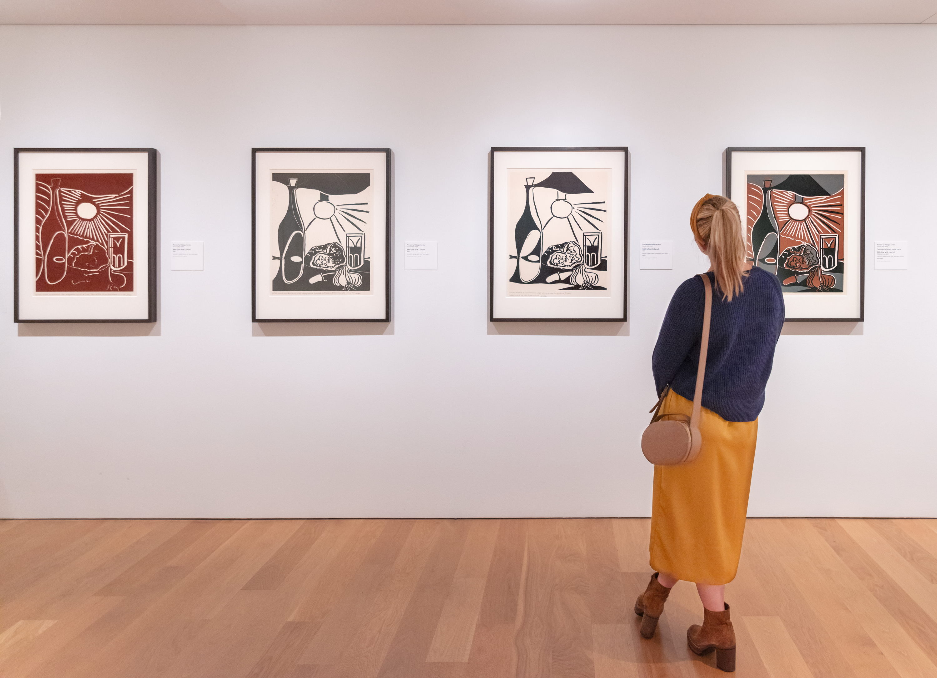 A blond woman with her back to the viewer stands facing a white wall on which hangs four versions of Picasso's print Still Life with Lunch, one white and dark brown-red, two in the center that are black and white, and on the right that is black, white, gray, and red-brown. The print image consists of a wine bottle, a lamp, a cut ham, a glass, a cut onion, and a knife.