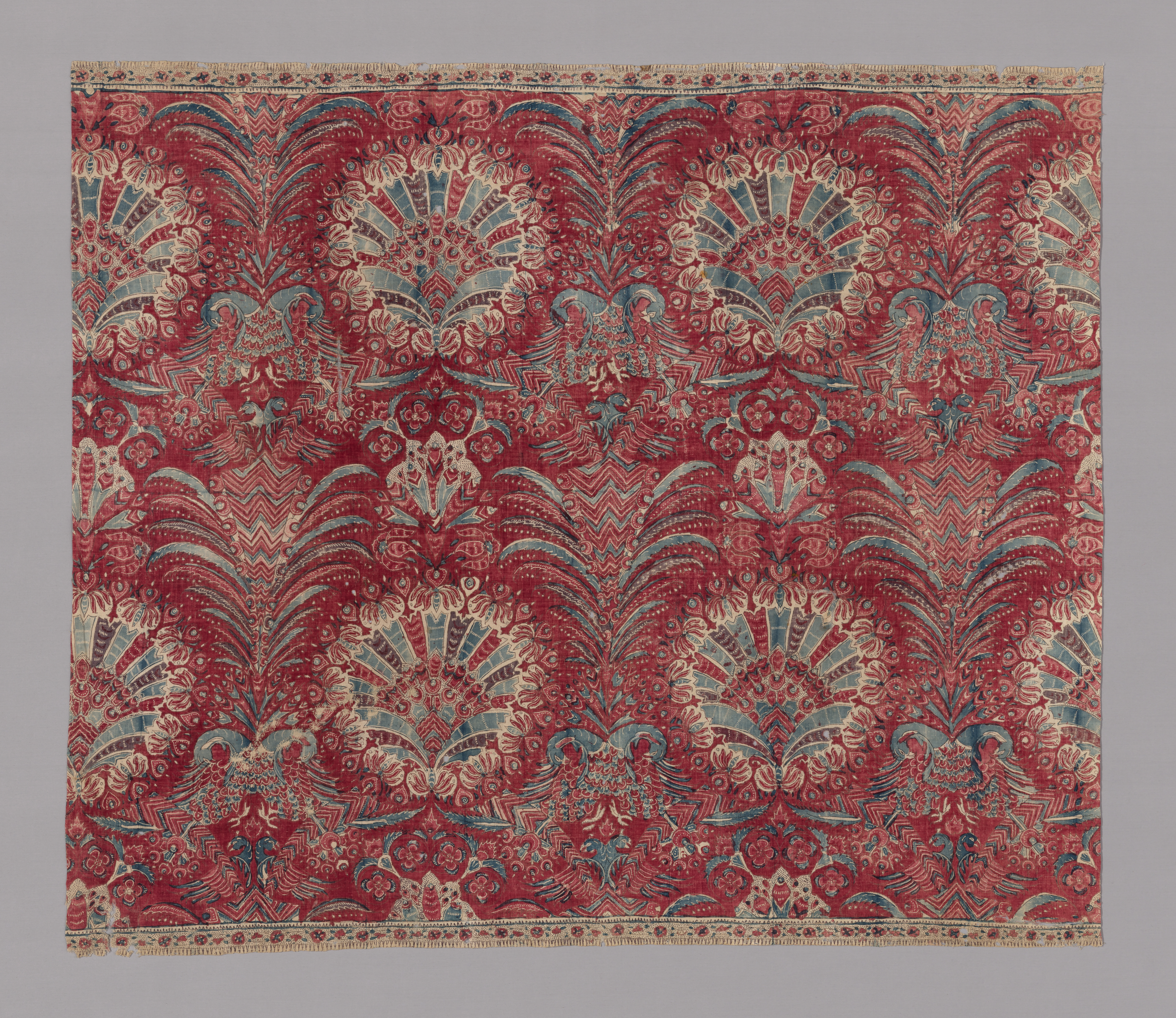 A swath of red fabric with a pattern in beige and light blue featuring foliage and flowers along with feather-like plumes and birds.