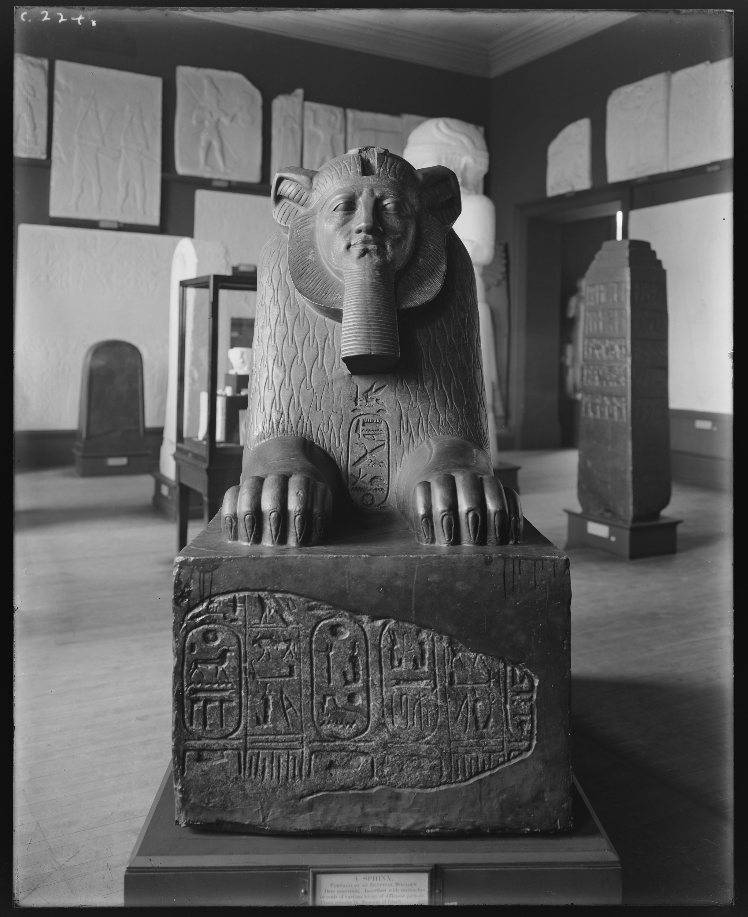 Black-and-white photograph shows the frontal view of a sphinx mounted on a carved pedestal.
