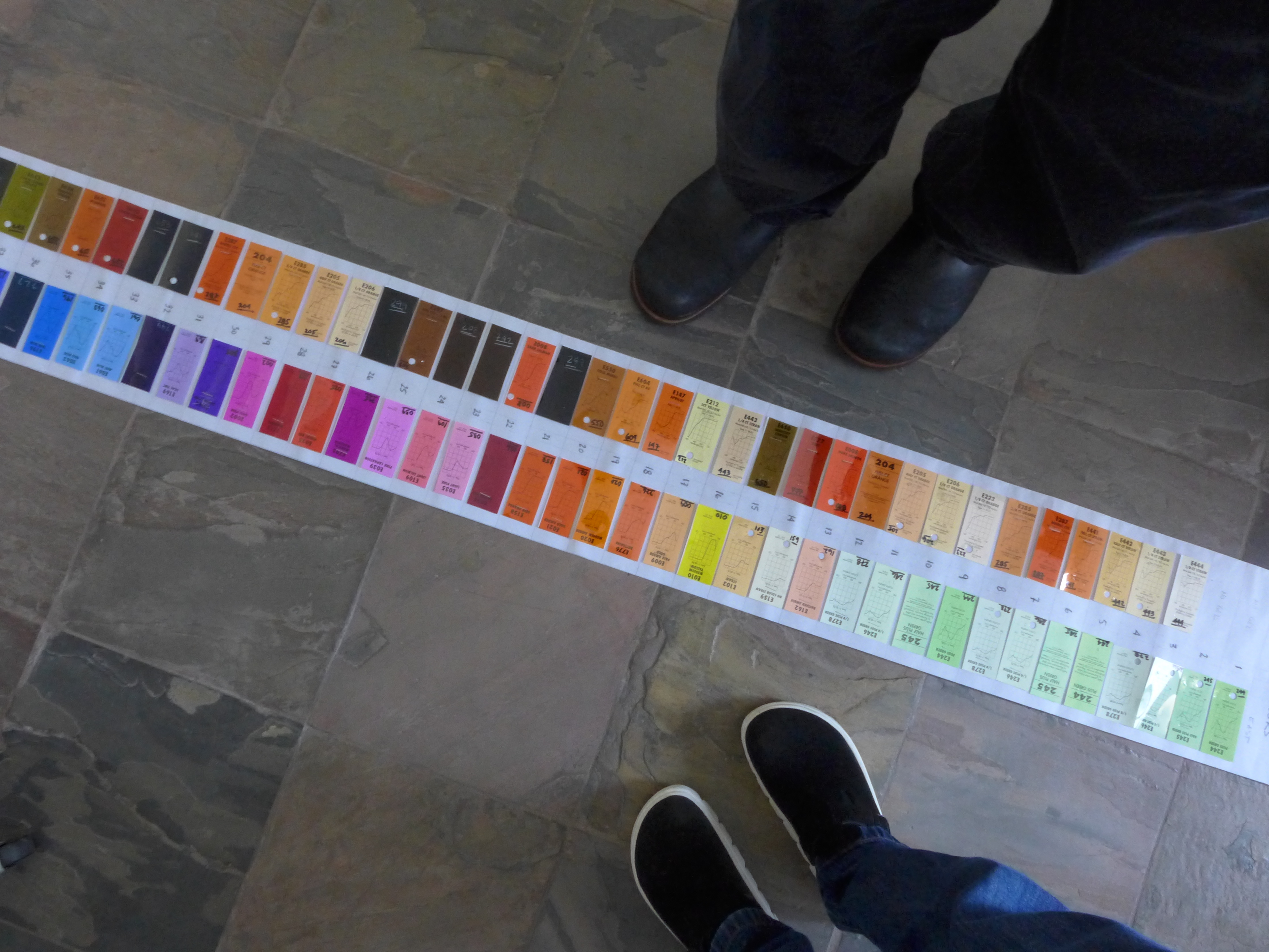 Photograph of a long stip of color swatches in two rows, in shades of green, pink, and orange, lined up on a brown tile floor, a pair of feet in shoes on either side.