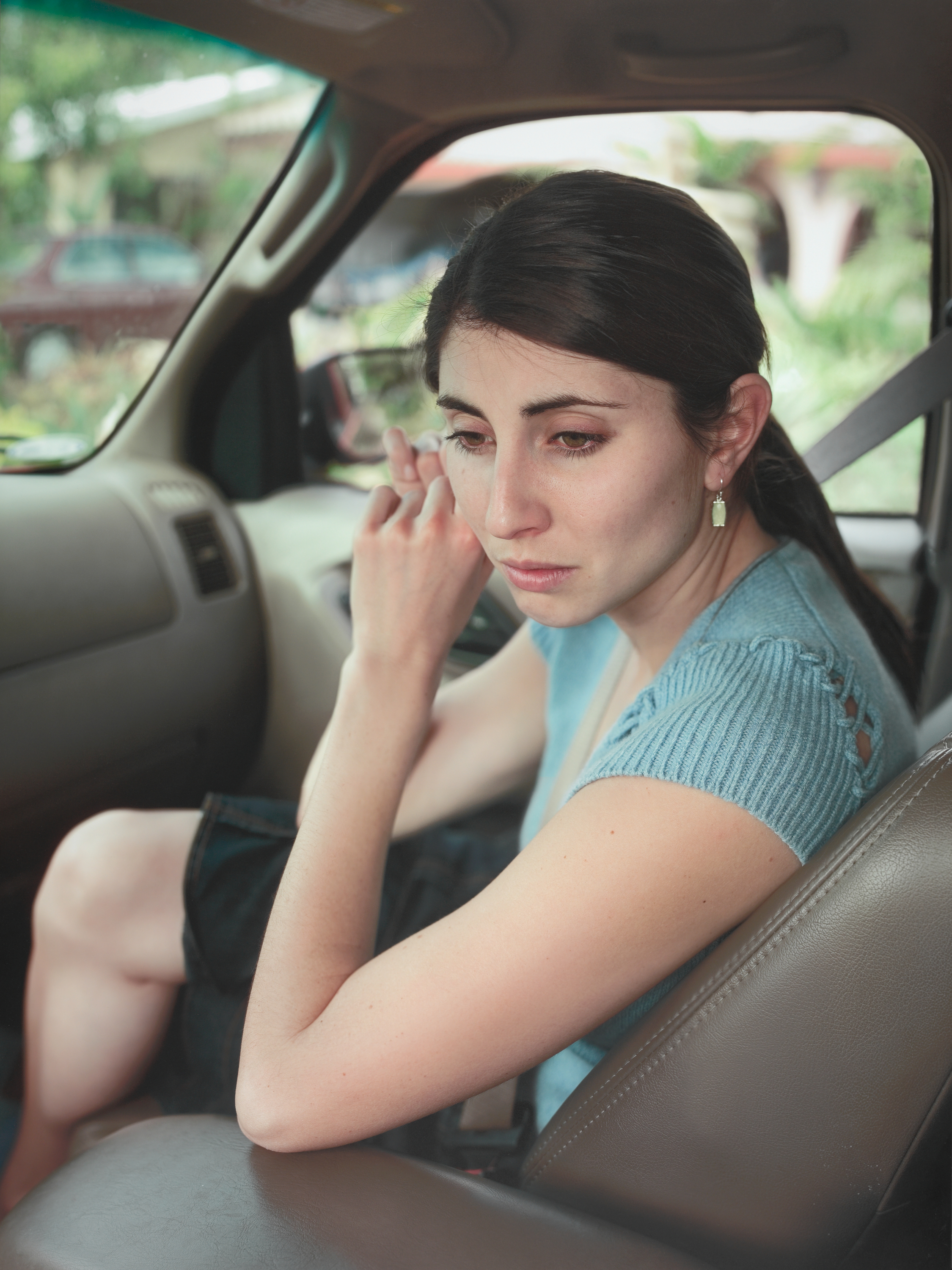 A color photograph of a young, light-skinned woman with dark hair sitting in a car with leather upholstery, fastening an earring while looking downward.