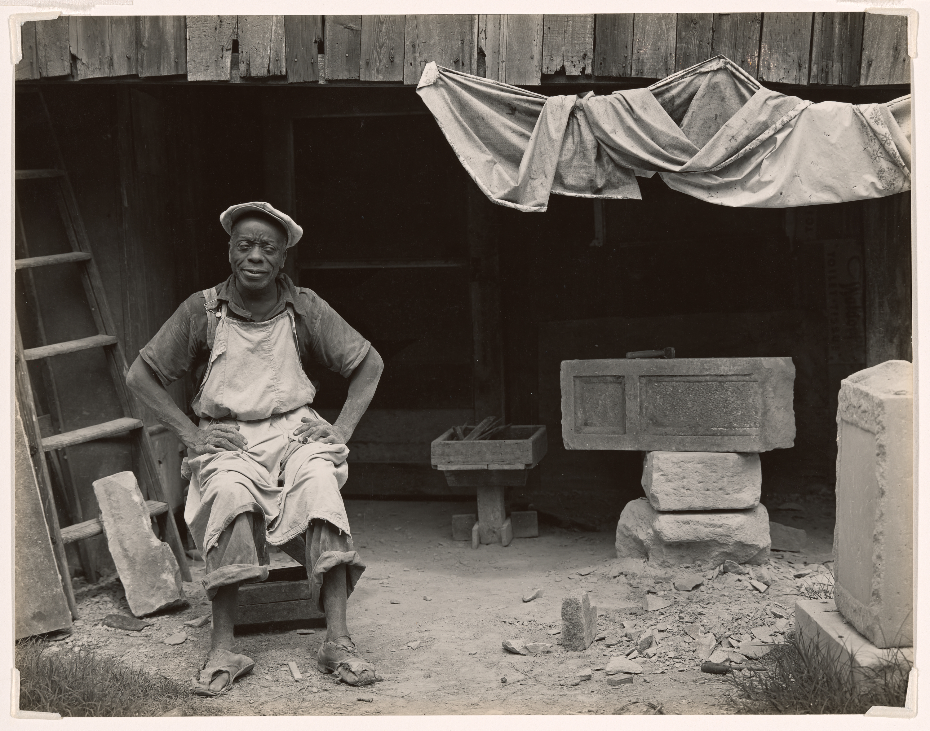 A black-and-white photograph shows an adult in a cap, shirt, apron, and rolled pants seated on a low stool at left-center with hands on hips and face obscured before a dark wooden shed; at left a ladder leans, at center a low table sits, and at right stone blocks and a basin rest under a draped cloth.