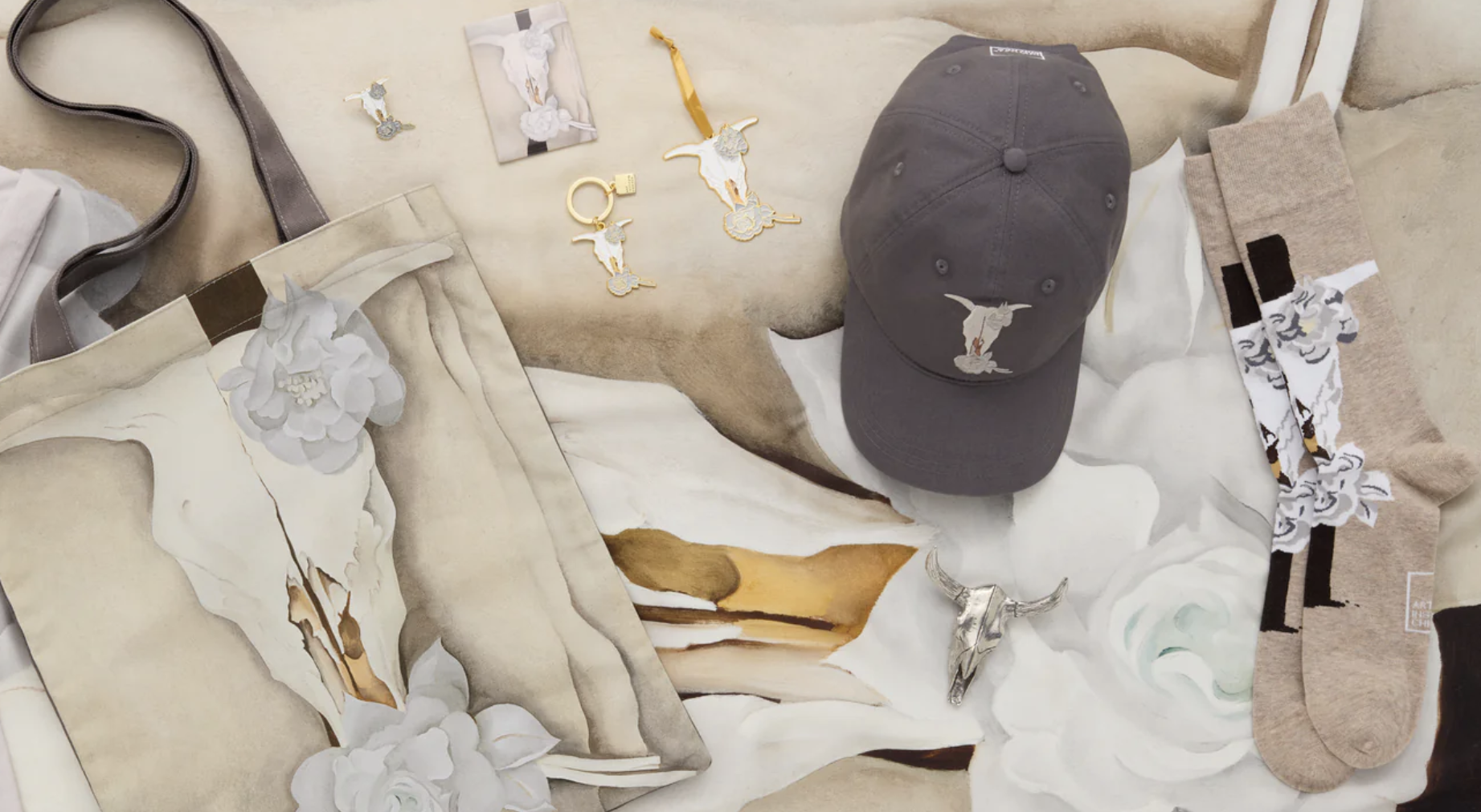Photograph of a wide variety of merchandise featuring a cow skull design by artist Georgia O'Keeffe. Among the items carefully laid out are a tote bag, a keychain, and a gray cap.