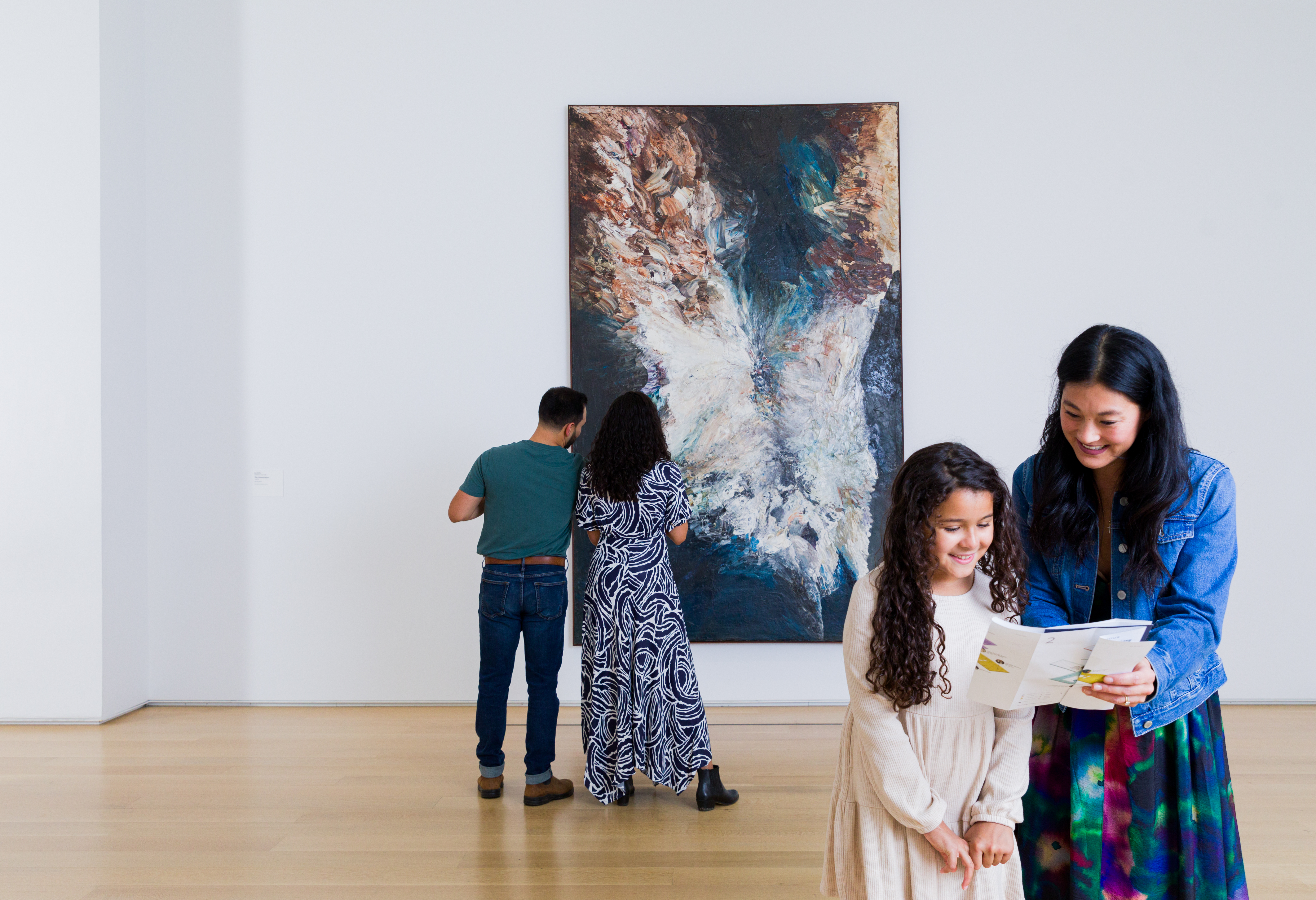 Photo shows a dark-haired young woman and girl in the foreground of a white gallery space looking at a printout while a young couple stands behind them, their backs to the viewer, looking at Jay DeFeo (Mary Joan DeFeo)’s The Annunciation, a large abstract painting suggesting large white wings with purple and teal on a black background.