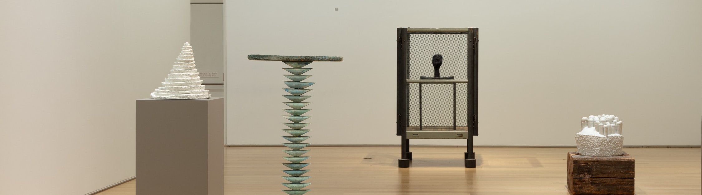 Photo of a white-walled art gallery containing six sculptures seen at a distance, each very different from the other: a white triangular form; a tall rectangular form; a thin, green rodlike form with a flat surface on top; a squat white form; and a globular red form that roughly evokes a female body.