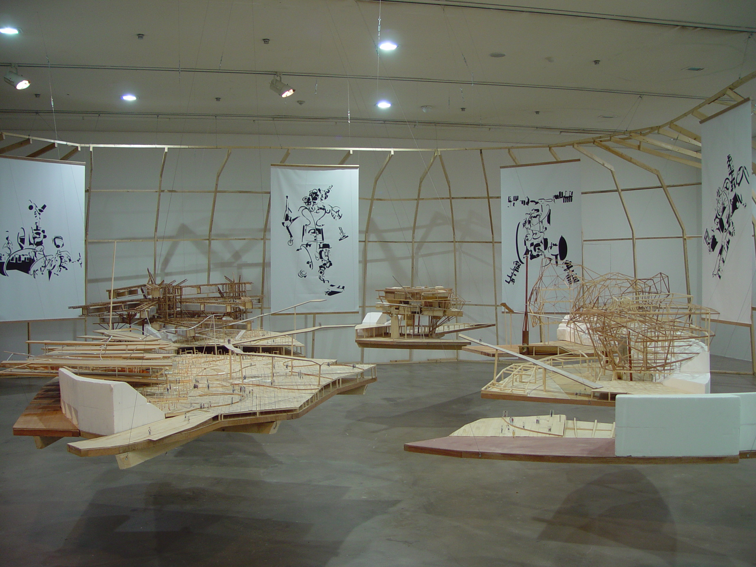 In a large room with concrete flooring, several floating models made of cardboard, tape, and string are suspended in the air. Four white banners with abstract drawings in black ink are suspended behind the models. Behind the banners, a large wooden structure surrounds the perimeter of the floating models and banners.