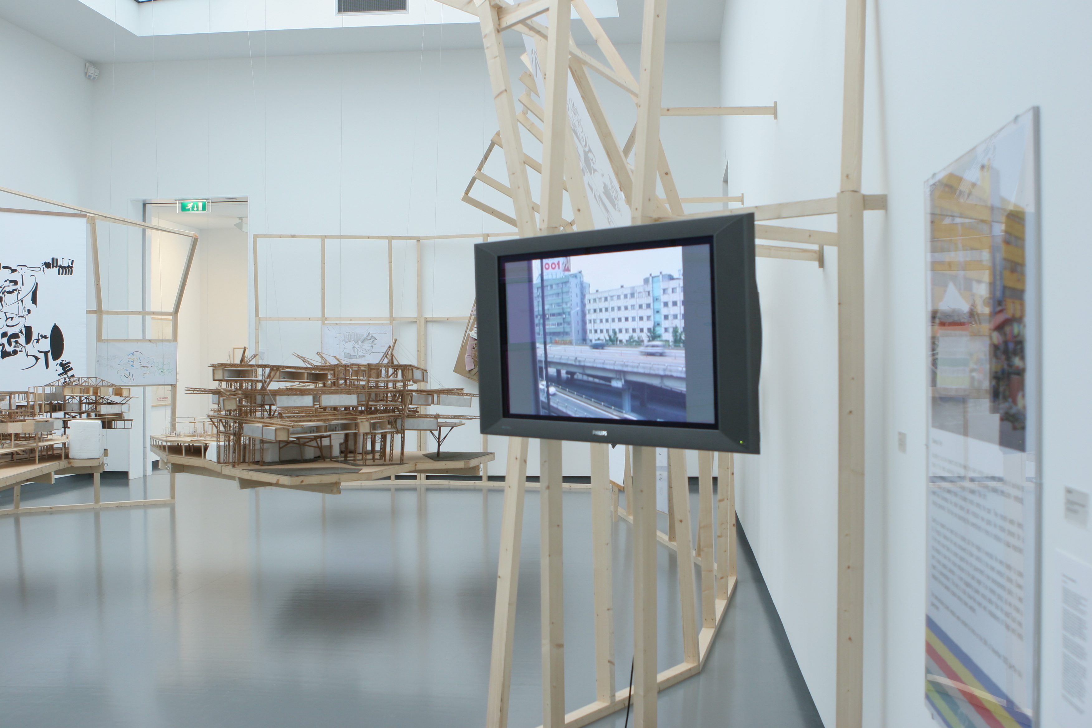 In a room with white walls, an LCD television showing a video of two cars driving along a ramp in an urban area is mounted on several long pieces of plywood. Behind the television, two models made of cardboard, wood, and string are suspended in the air, alongside a white banner displaying an abstract drawing in black ink. To the right of the television, a gallery label displays information on the installation.