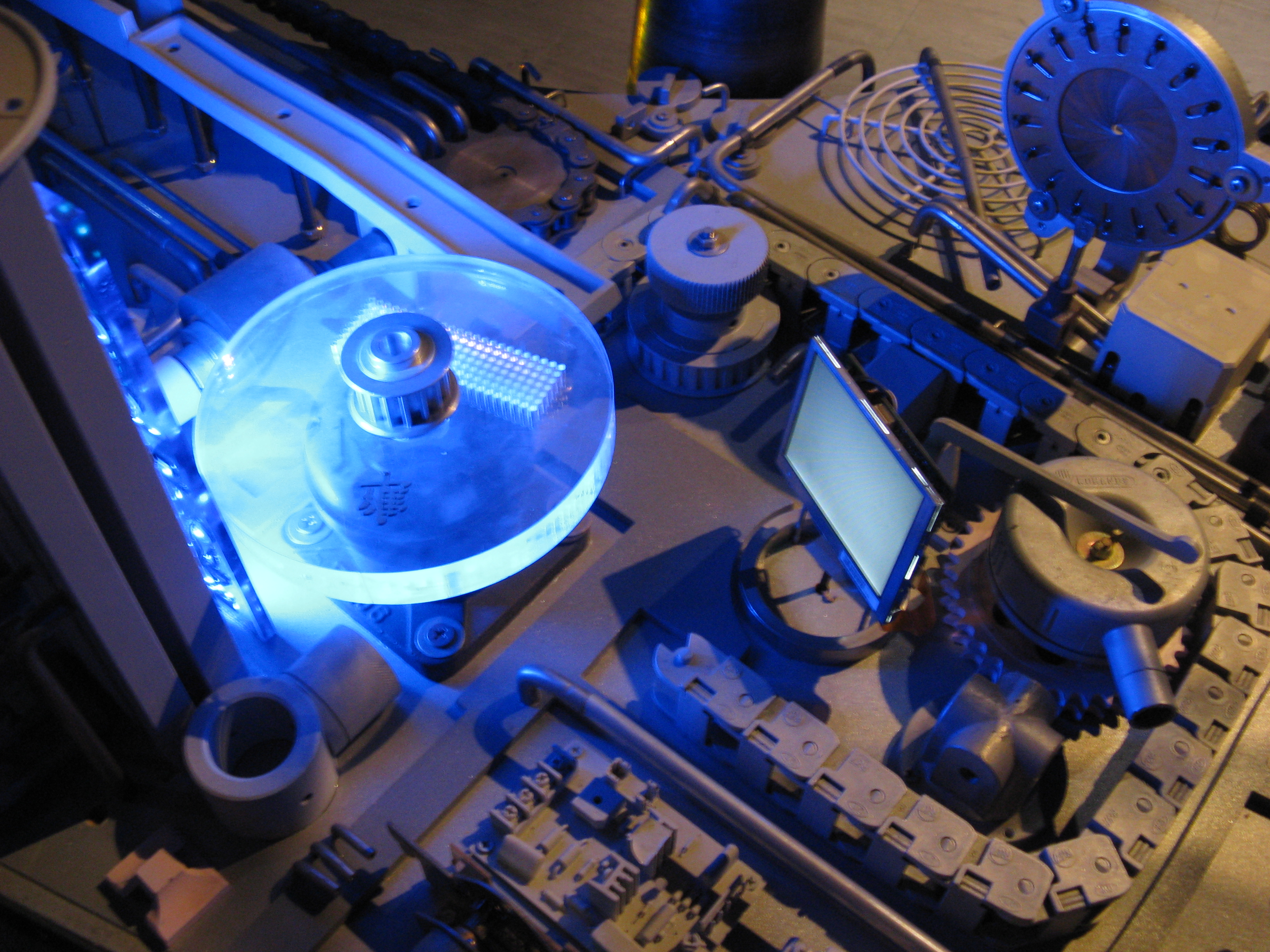 Several cogs, rotors, mechanical chains, and other mechanical parts rest on a board. The rotor at left is made of a clear material, and has vivid blue light reflected on it by a spotlight.