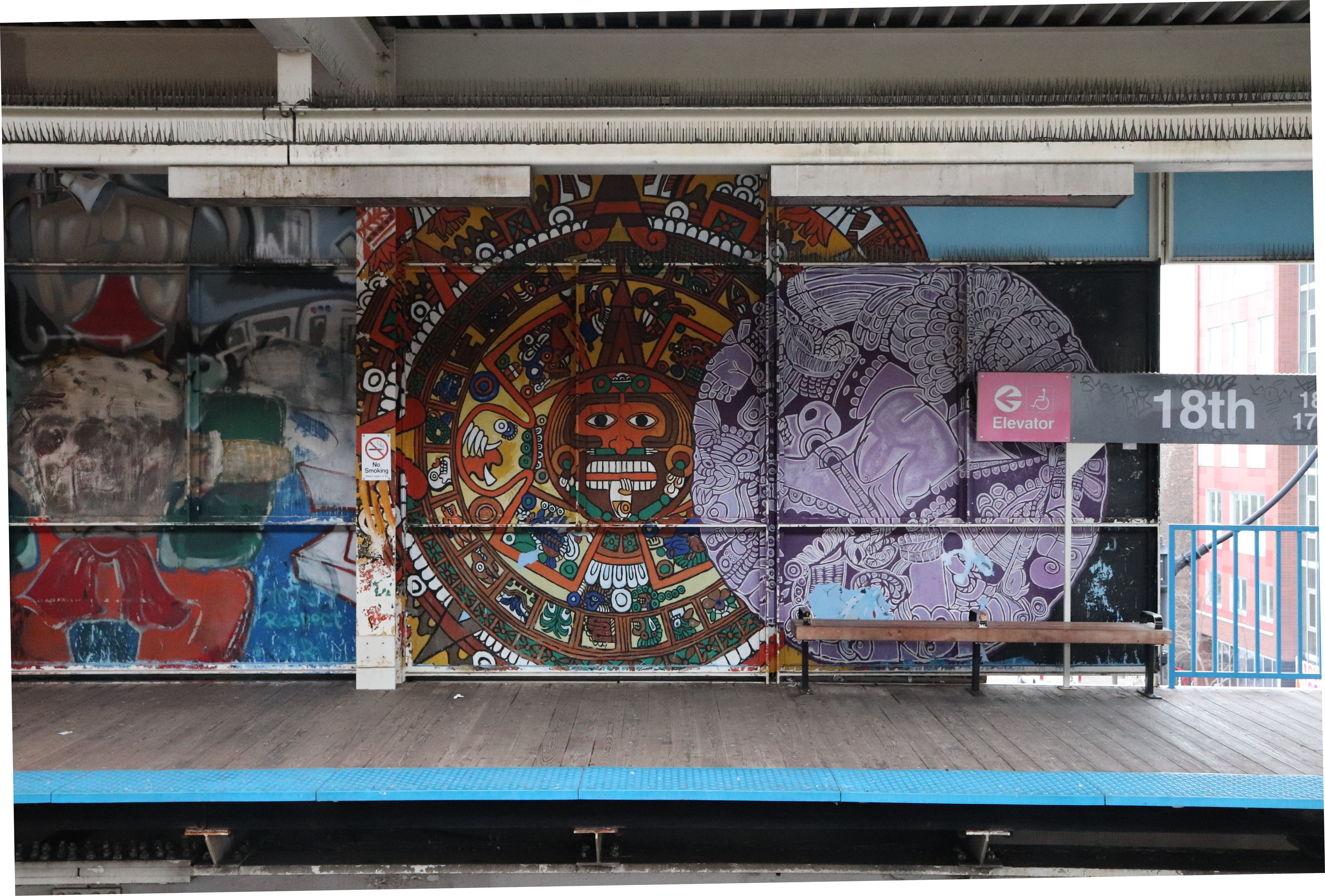 A color photograph of an elevated train platform shows a large mural; at center a circular orange face with red, green, and yellow geometric patterns, at right a purple patterned circle and a brown bench below a gray 18th and pink Elevator sign, with mixed graffiti at left.