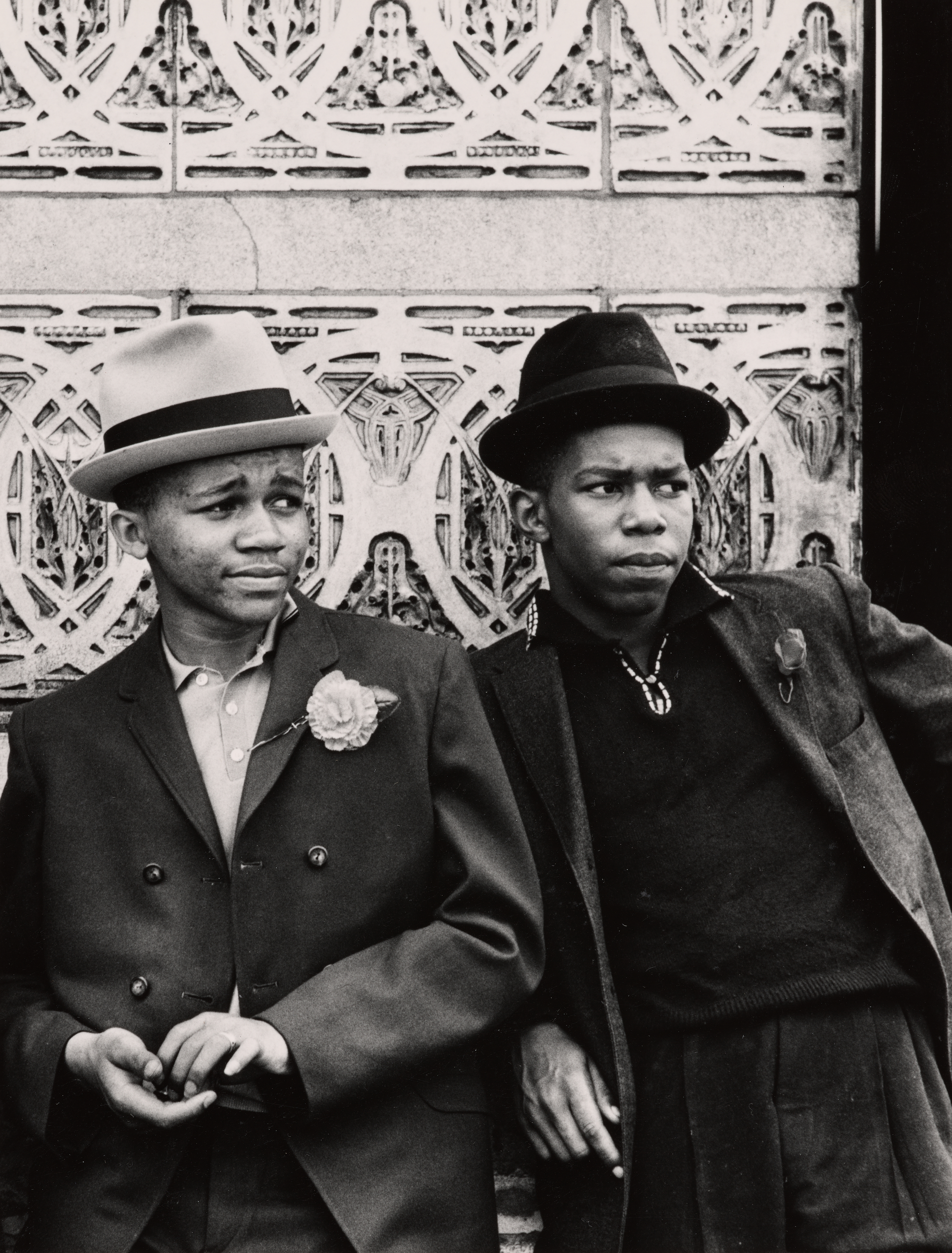 Black-and-white photograph of two young men standing in front of a decorative, patterned stone wall. Both wear dark suits and brimmed hats. The man on the left has a flower pinned to his lapel and a relaxed expression, while the man on the right wears a necklace and has a more serious gaze. They lean casually against the wall, with their bodies slightly angled toward each other.