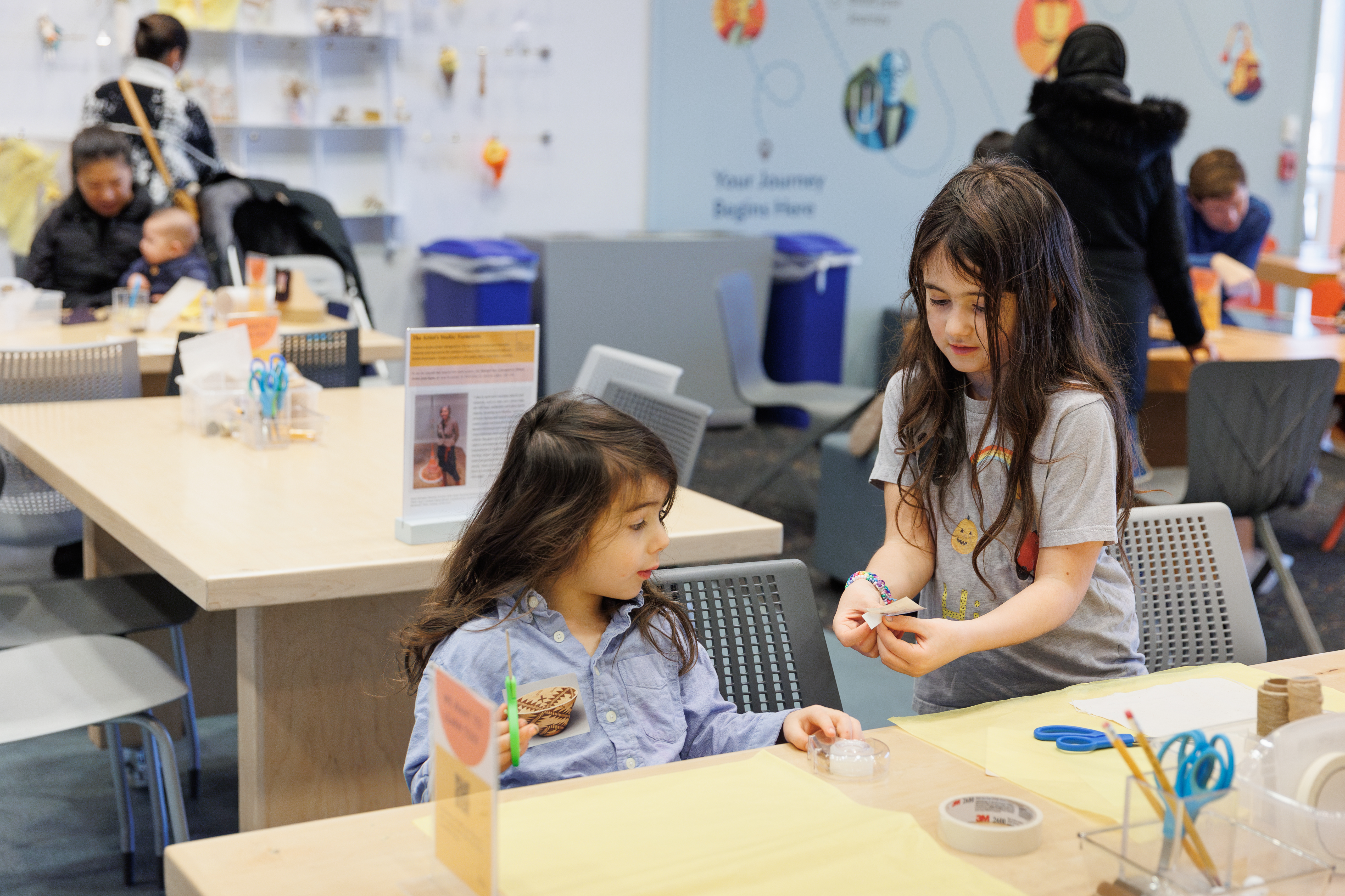 A photograph shows two young girls with light skin and dark hair at a table with pencils and scissors. The older girl, at right, stands and hands the younger girl a bit of folded paper as the young girl looks on with interest.