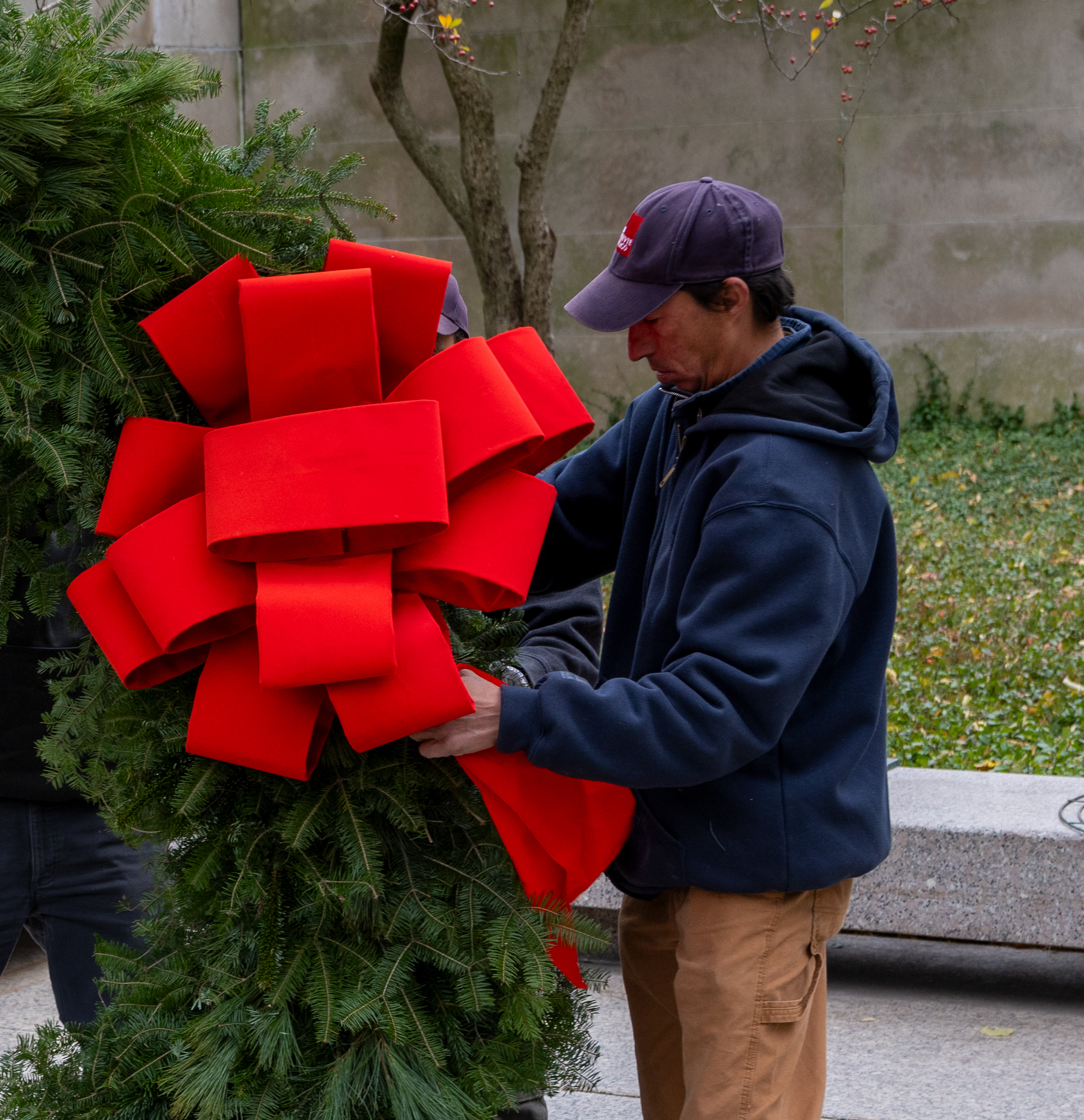 In a photo, a slim man with medium-colored skin affixes a large, red, velvet bow to a large evergreen wreath.