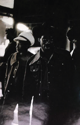 Photograph of three Black men in shadowed indoor light, walking closely together. The central figure wears a dark cowboy hat and denim jacket with white stitching, partially illuminated across the chest. The man to the left wears a lighter-colored hat and a coat with a wide collar, while the third man in the background wears a brimmed hat. Bright reflections appear at the bottom edge of the image.