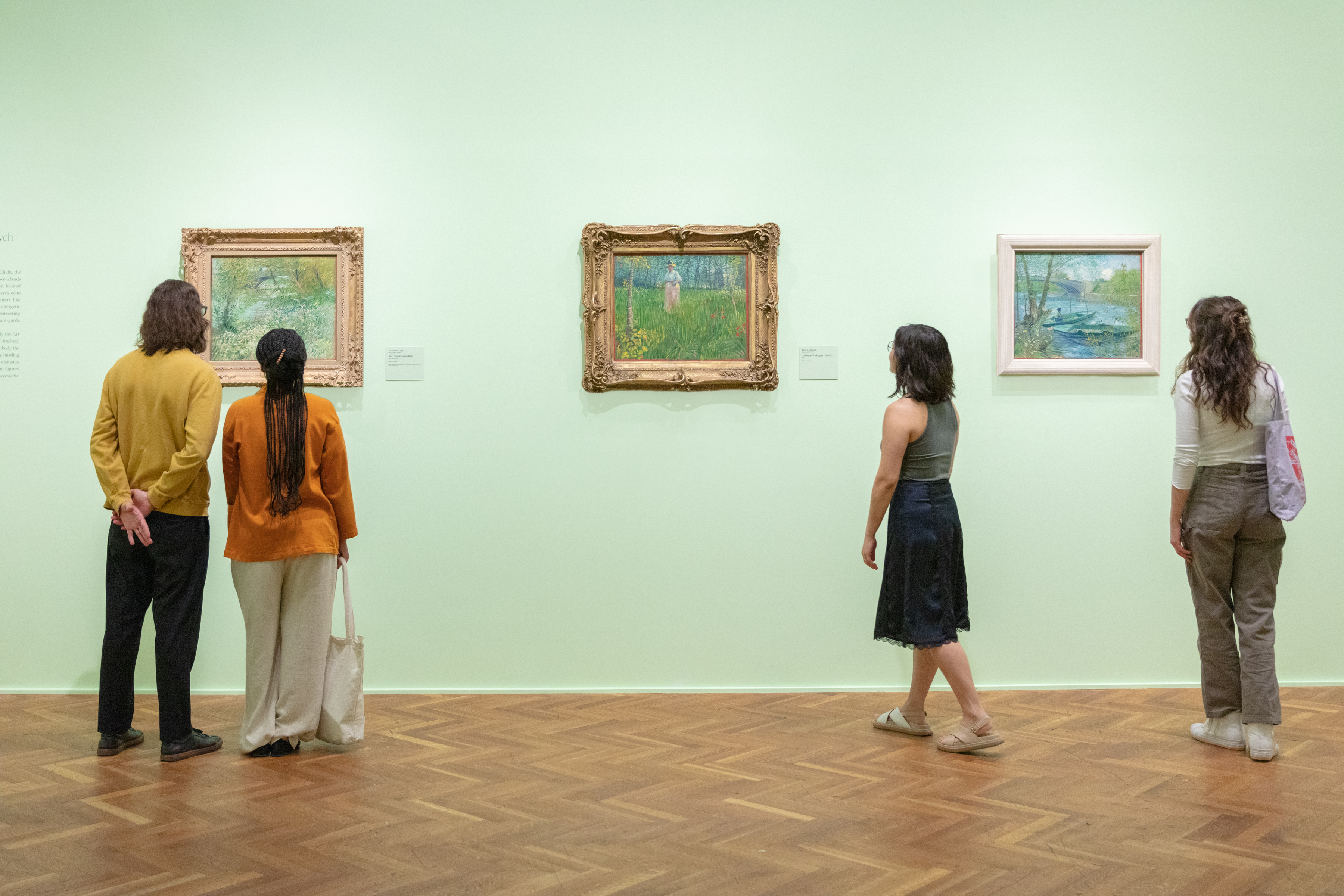 Photograph of a mint-green art gallery in which several young adults, their bacs to the viewer, closely observe three separate works by Vincent van Gogh: "River Bank in Springtime" at left, "A Woman Walking in a Garden" at center, and "Fishing in Spring, the Pont de Clichy (Asnières)" at right.
