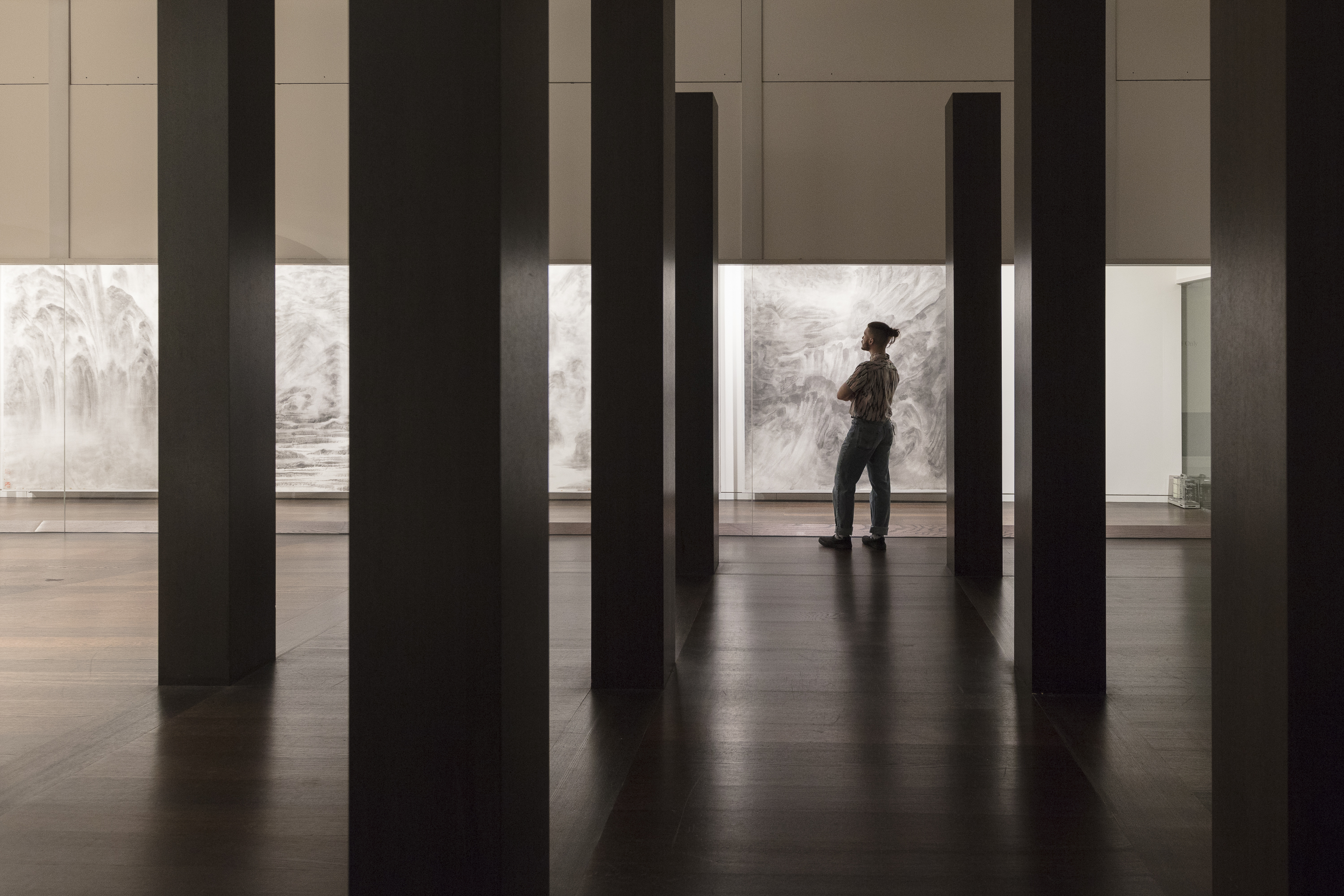 A man stands between rows of tall, rectangular, black columns looking at a series of grayscale landscape paintings.