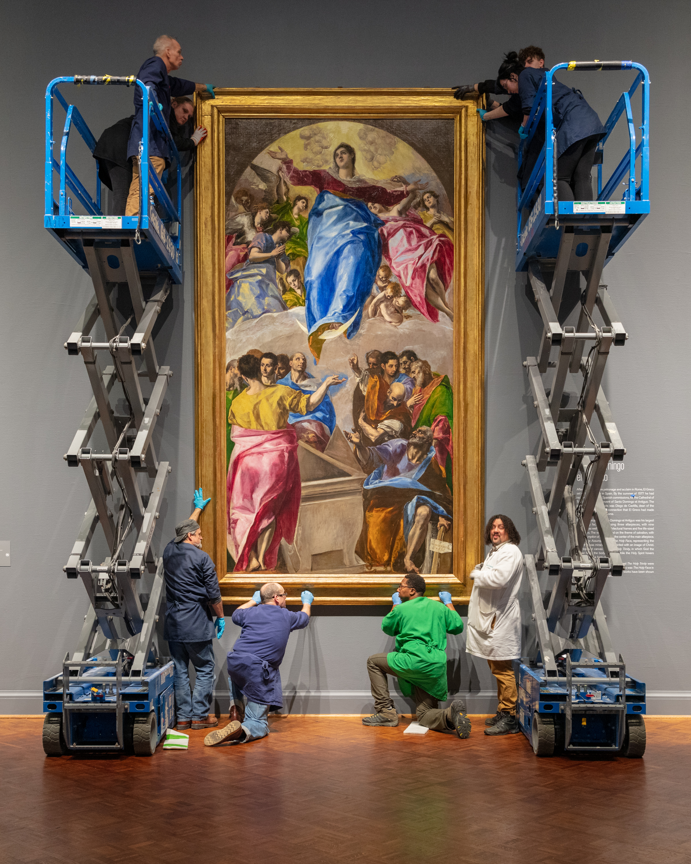 A group of workers in gloves and aprons install a large Renaissance painting, El Greco's The Assumption of the Virgin. Four people hold the bottom of the painting. On either side, four others use a mechanical lift to secure the top.