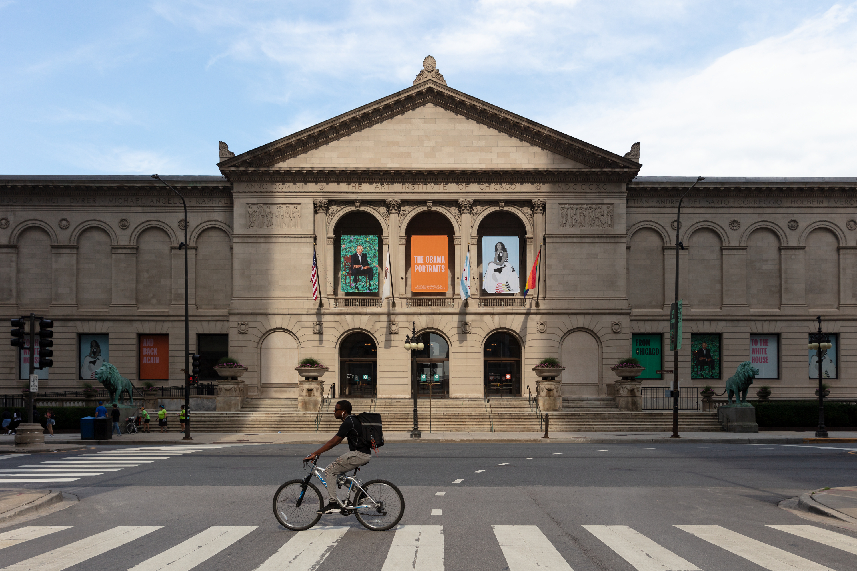A man rides his bike in front of the Art Institute of Chicago, a two-story, limestone building with a central portico, blank pediment, and symmetrical wings decorated with archways.