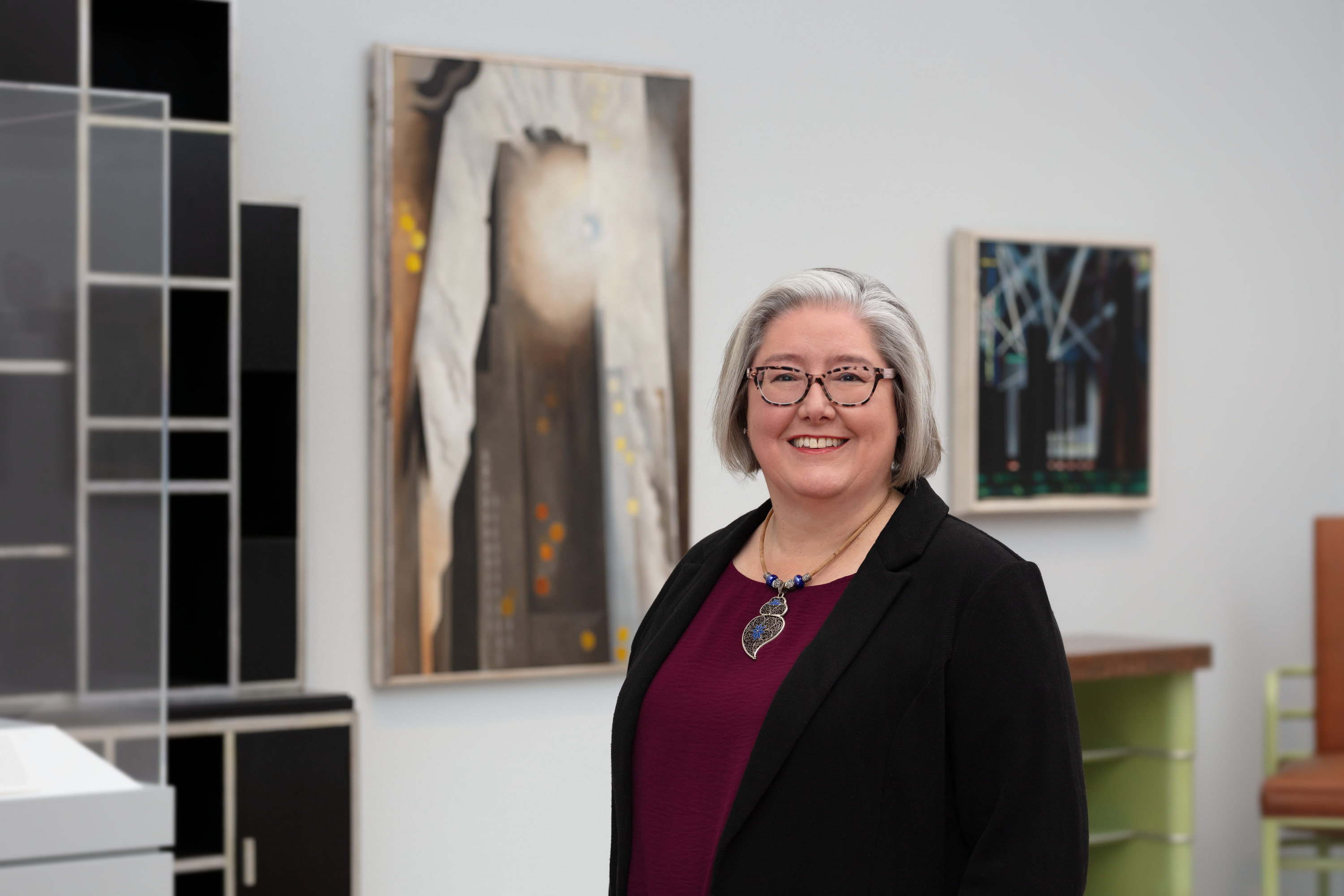 A light-skinned woman with gray and white hair, Sarah Kelly Oehler, stands smiling in a black jacket and burgundy shirt with Georgia O'Keeffe's painting "The Shelton with Sun Spots" behind her at left.