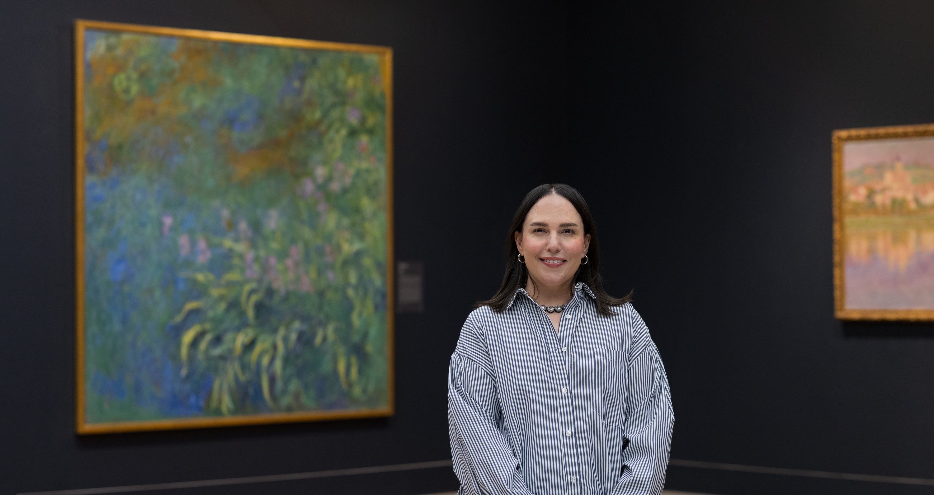 A young woman with long dark hair, Zahra Bahia, wears a striped shirt and stands, facing the viewer, before a display of three paintings of haystacks by Claude Monet.