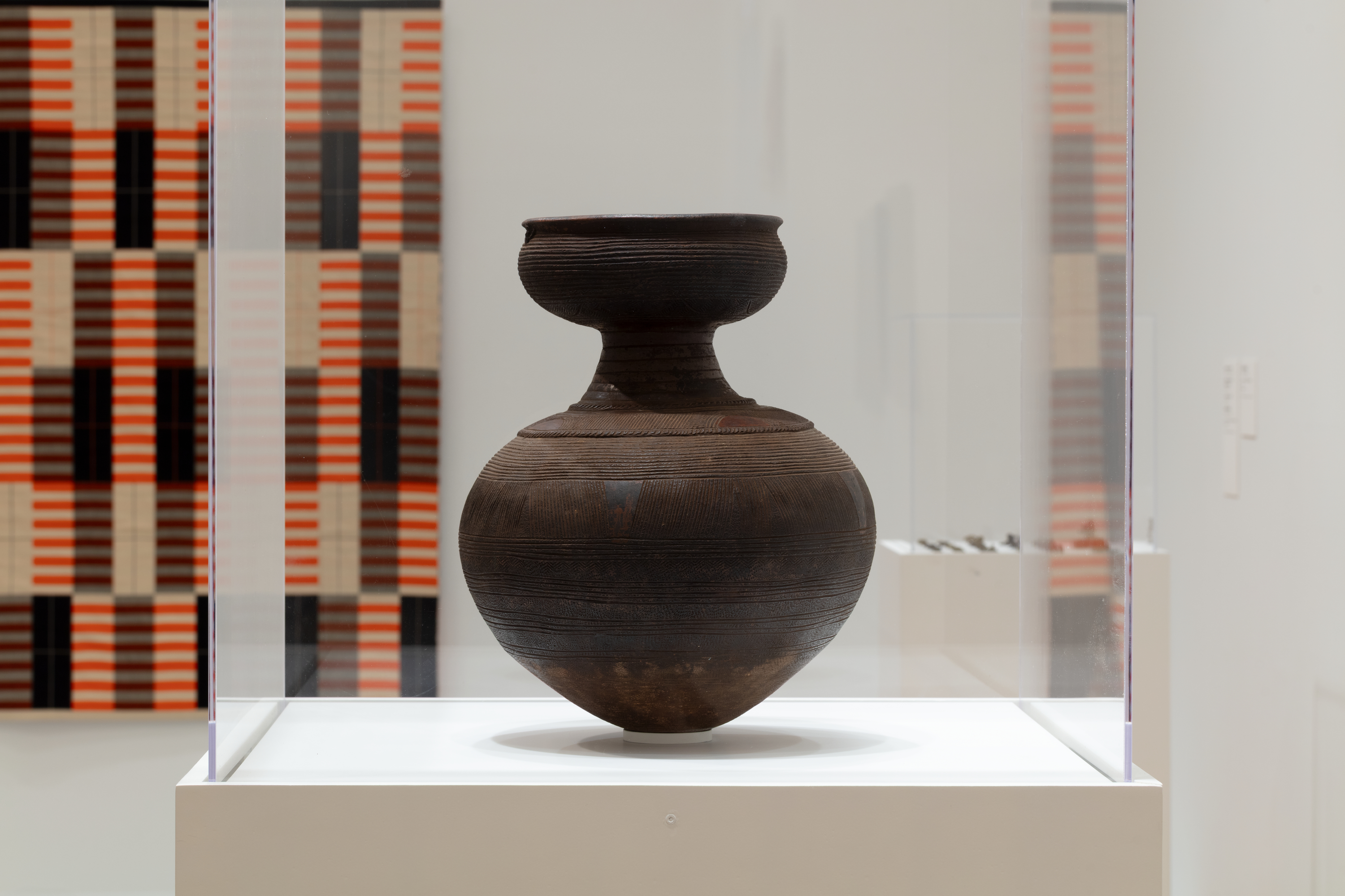 A brown ceramic pot in a plexiglass vitrine. The pot has a flat and wide opening at top that funnels to a narrow neck and opens again into a wide bulb and then narrowing to a tiny base.