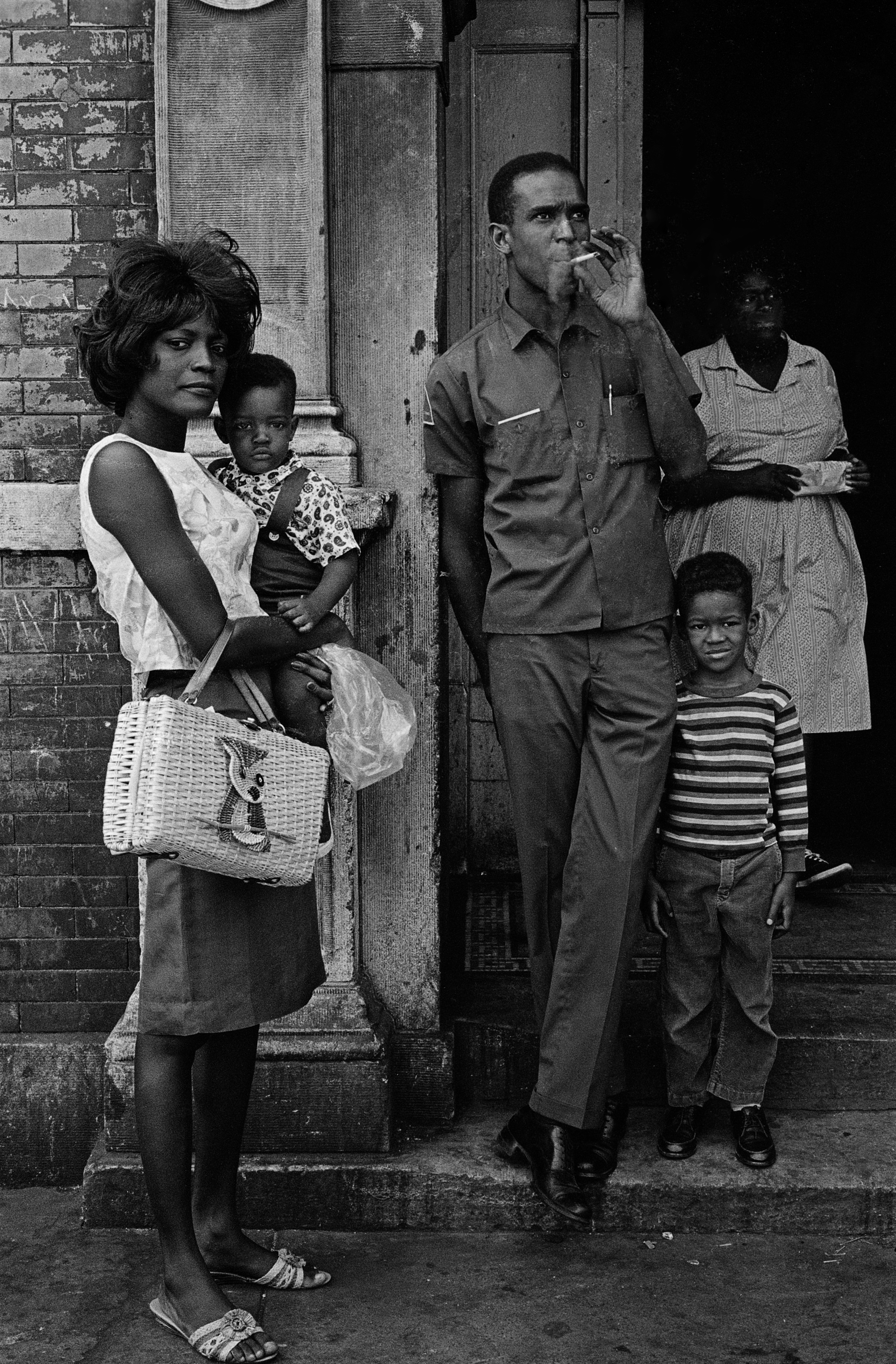 Black and white photograph of five people gathered in front of a brick building entrance. At left, a woman stands holding a small child on her hip; she wears a sleeveless blouse, skirt, open-toed sandals, and carries a woven purse with a duck motif. Beside her, a man in a uniform shirt smokes a cigarette, with one hand in his pocket. A boy in a striped shirt and cuffed pants stands in front of him, looking at the camera. In the shadowy doorway behind them, a woman in a patterned dress holds a dish. The scene appears casual yet composed, with all subjects either facing or aware of the camera.