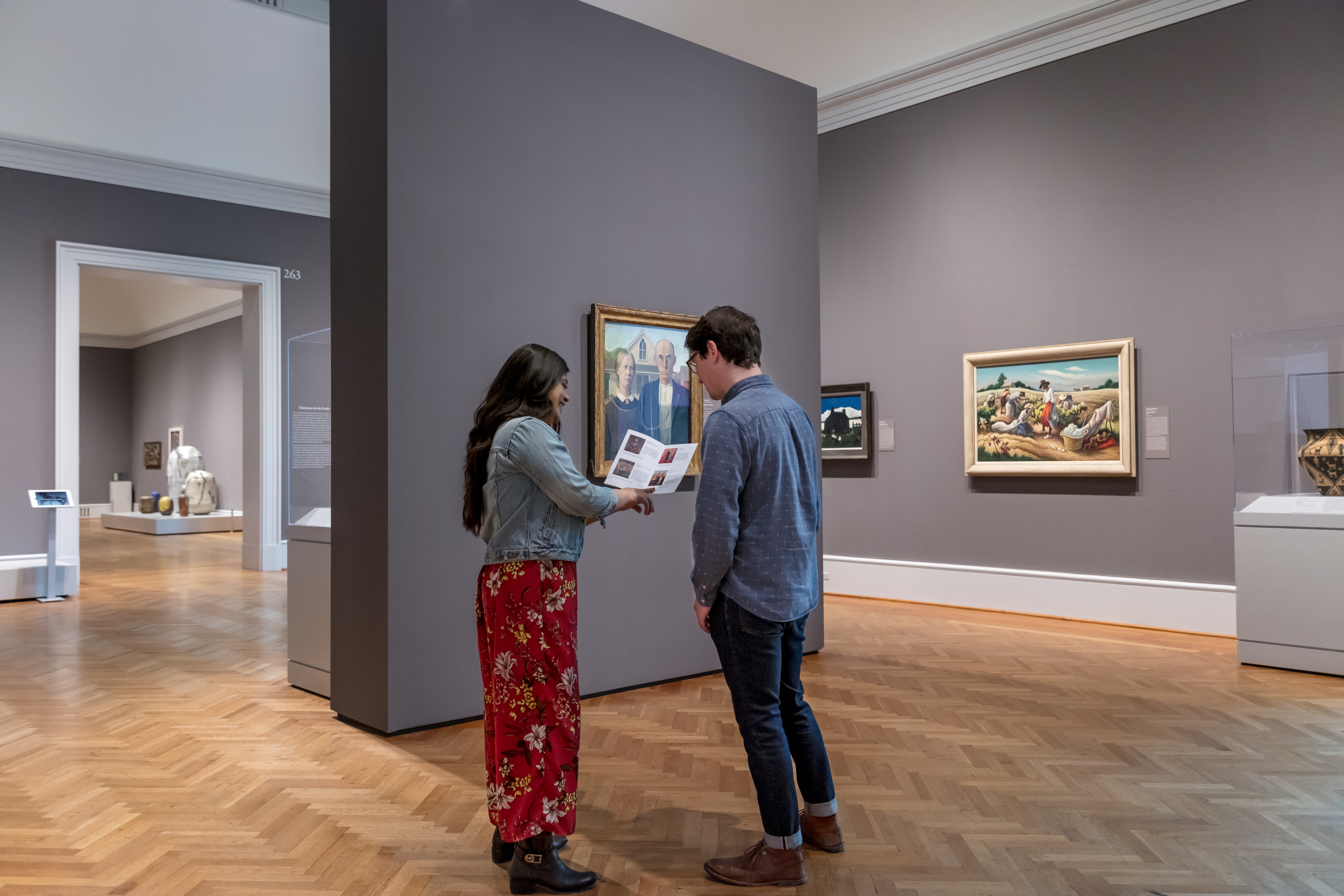 A medium-skinned woman with long black hair points to an image in a folded pamphlet while standing with a light-skinned man in front of Grant Wood's painting American Gothic. An array of other artworks fill the galleries surrounding them.