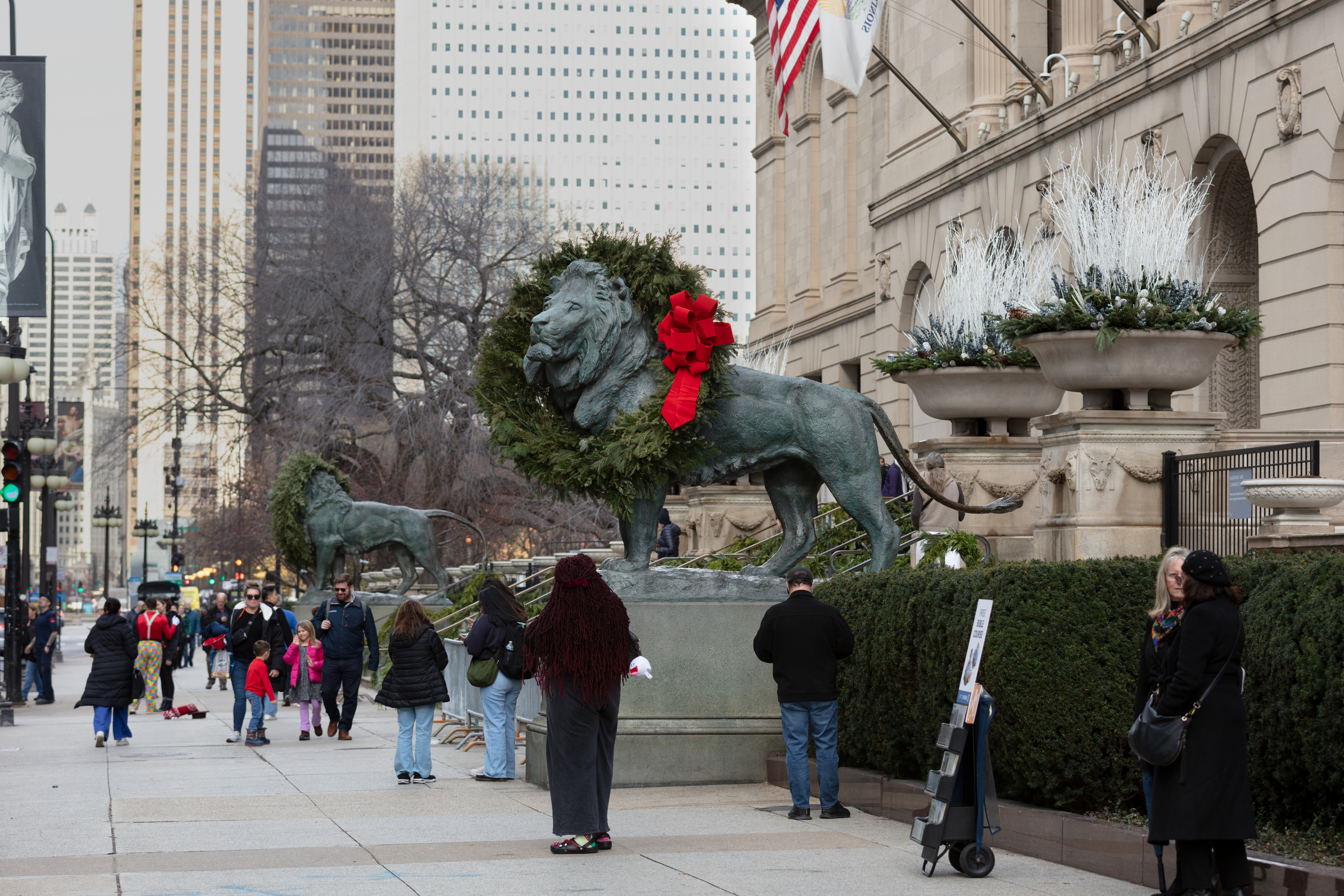 A photo taken outside the Art Institute of Chicago shows two large bronze lion sculptures adorned with evergreen wreaths. People bustle by on the sidewalk around them.