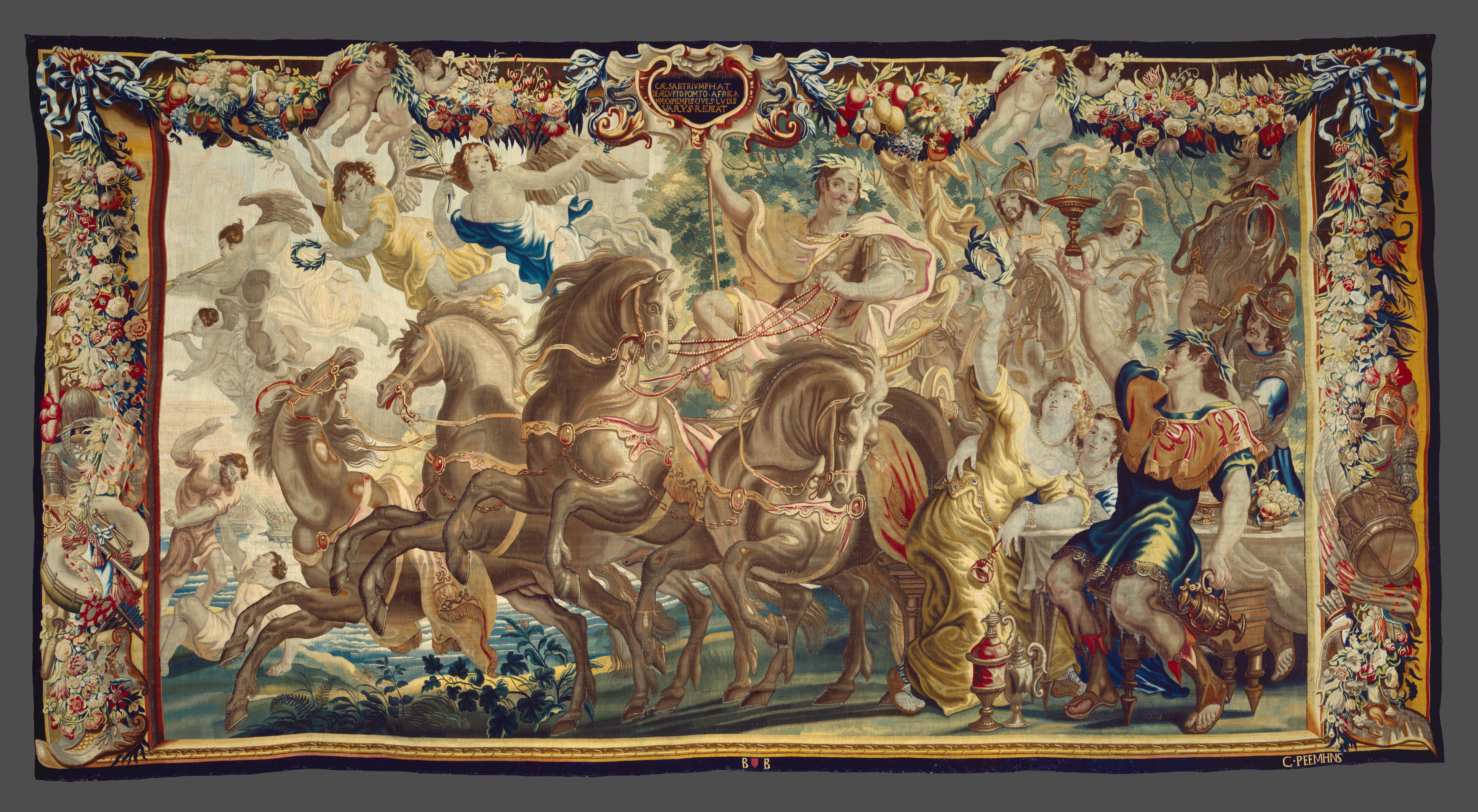 A large tapestry with an ornate border featuring a man seated on a charriot drawn by four horses passing by a table of revelers.