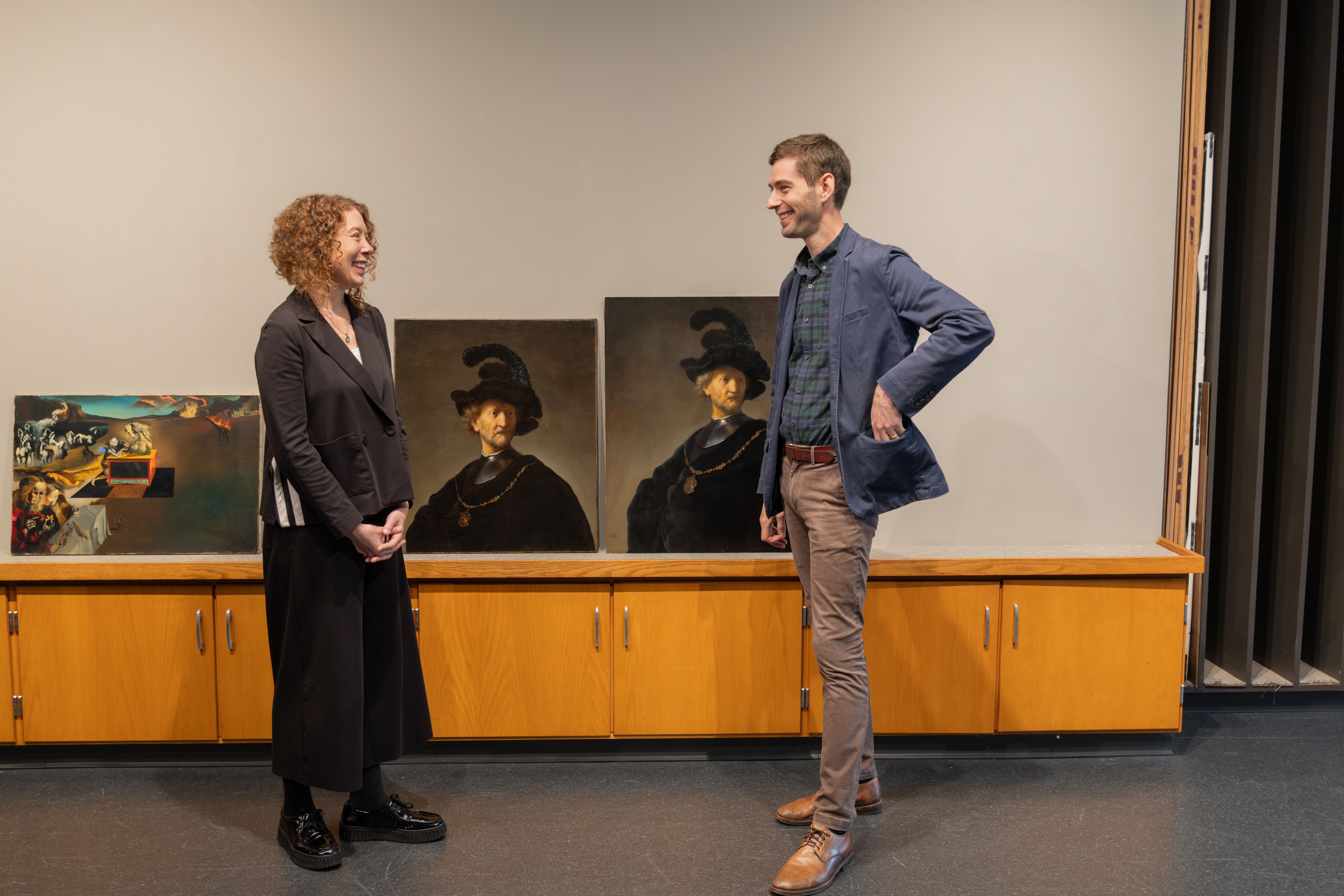A light-skinned woman with curly reddish hair, Jacquelyn N. Coutré, and a tall, light-skinned man with light-brown hair, Gerrit Albertson, face each other smiling in profile. Between them are two paintings of the same subject: an old man wearing black and a feathered hat. The painting on the right is slightly larger than the one at left.