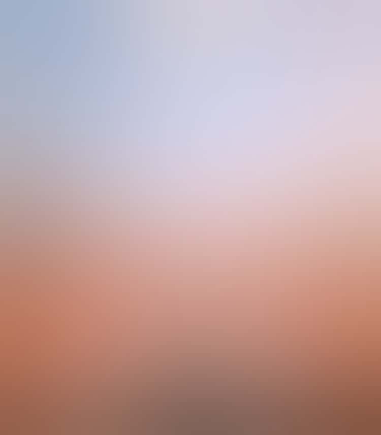 A blistering, halo-like sun shines strongly down through a wide, blue sky to a gray and hazy cityscape of industrial buildings and a river illuminated in red, smoke rising from buildings and smokestacks.