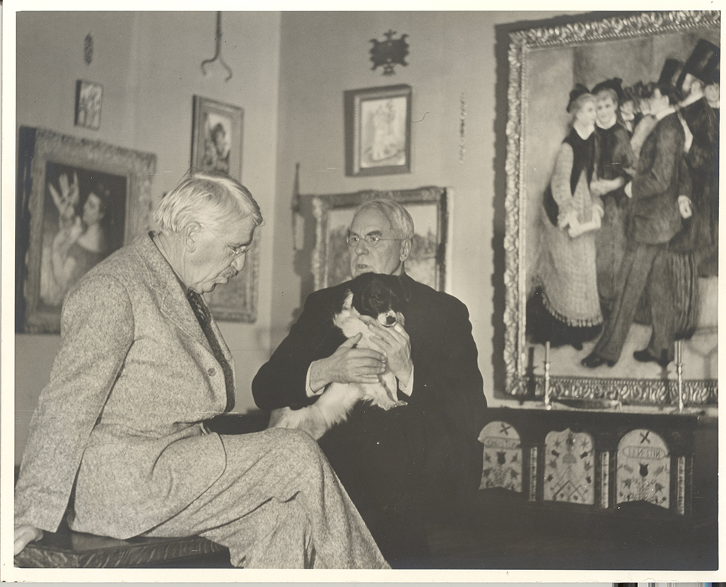 Top to bottom, the back wall holds several framed pictures; a very large frame on the right shows a group of standing figures, while smaller frames to the left and center show single figures. In front of them, two older adults sit close together; the left person wears a light gray suit, and the right person wears a dark suit and holds a small, long‑haired dog that is mostly white with dark patches. Along the lower right, a low cabinet or bench with patterned panels sits beneath the large frame, with two candlesticks placed in front of it.