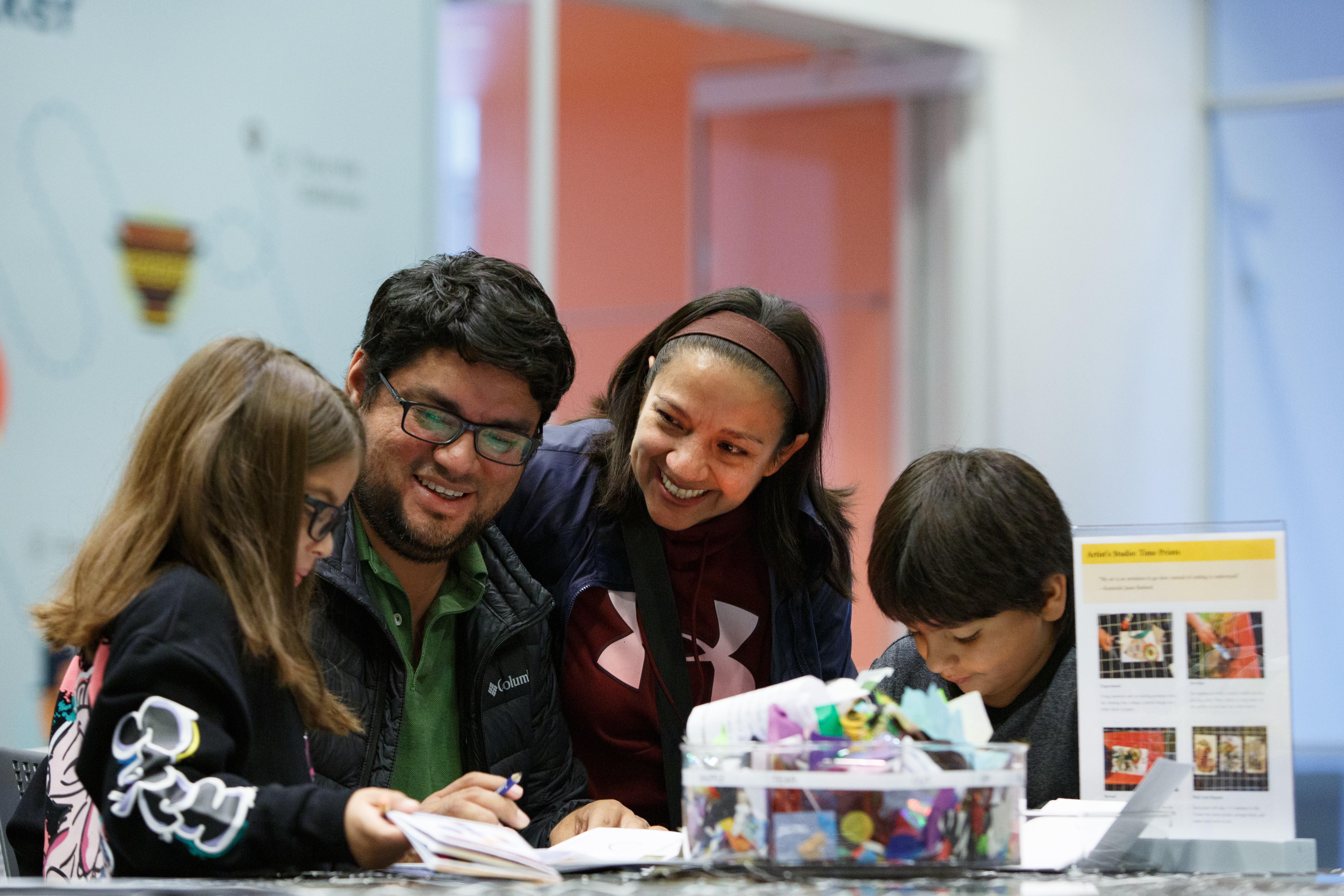 A photo shows a smiling family of four people with medium skin—a man, woman, young girl, and young boy—at a table creating artwork together.