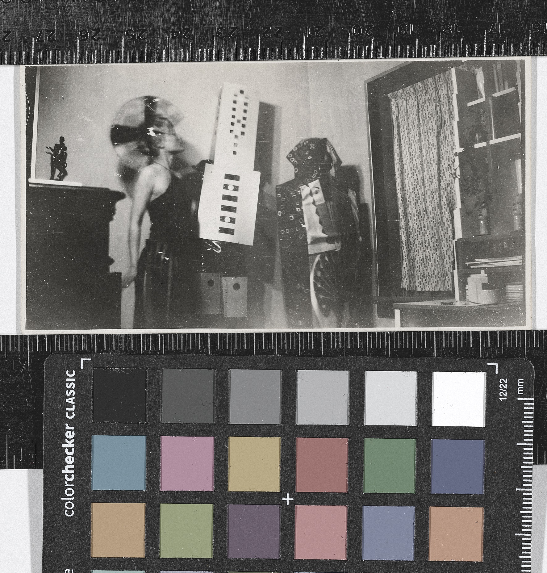 A black-and-white photograph of three costumed people in an interior home-like space. A woman wears a black one-shouldered dress with an elaborate headdress. The person in the middle wears a structure that looks like a skyscraper covering their head and torso, and the person on the right is draped in panels of what appears to be textiles with different images and patterns.