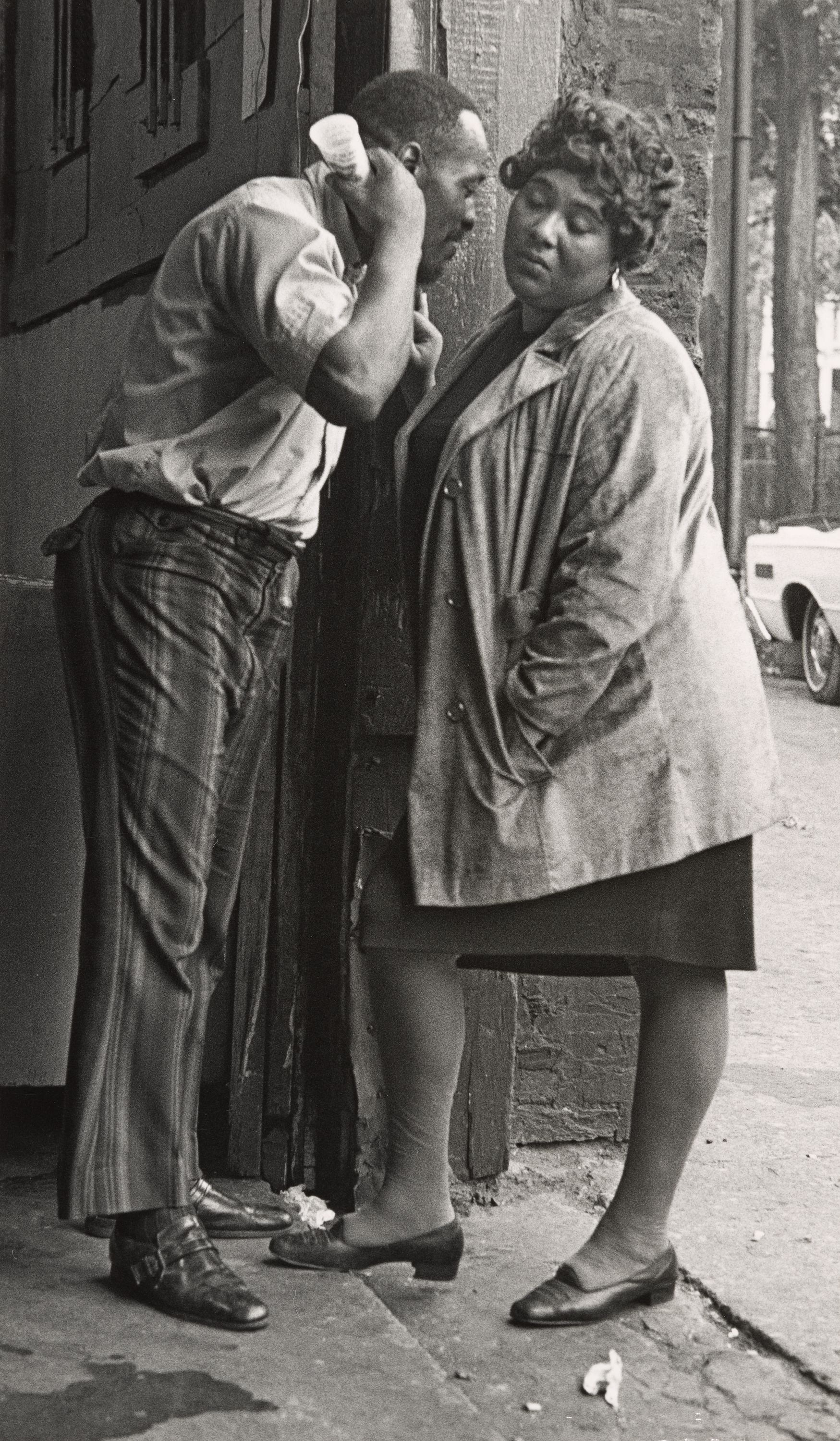 Black-and-white photograph of a man and woman standing close together outside a brick building. The man leans in with one hand raised behind his head and a small cup in the other, as if whispering or speaking quietly to the woman. He wears a short-sleeved shirt and striped trousers. The woman, wearing a dress and long coat, stands with her eyes closed and head tilted slightly downward. A parked car and tree trunks are visible in the background along the street.