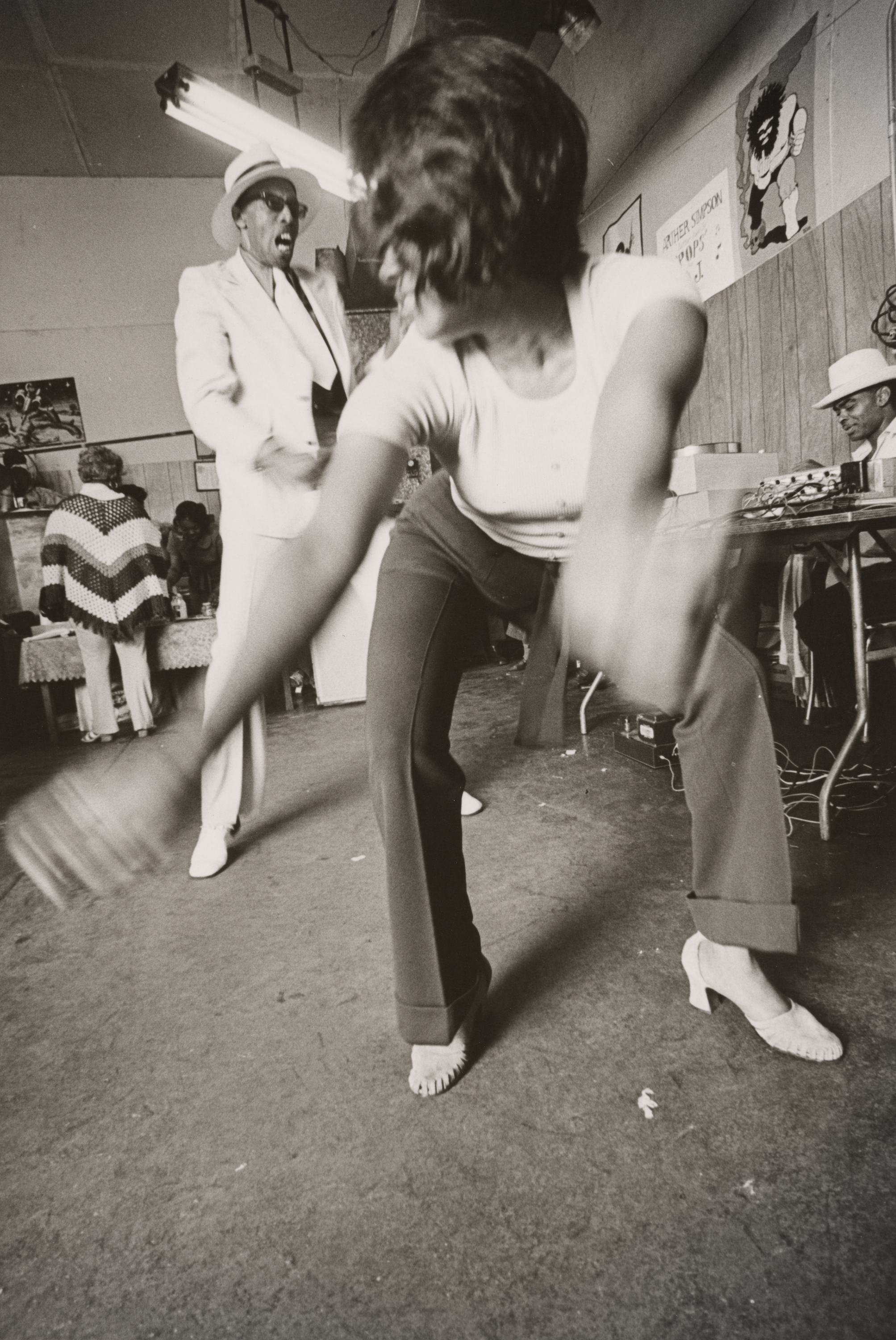 Black-and-white photograph of a woman dancing in the foreground, bent forward with arms swinging wide and blurred in motion. She wears a short-sleeve top, flared pants, and heeled shoes. Behind her, a man in a white suit, dark shirt, and wide-brimmed hat appears to be singing or shouting with an animated expression. The scene takes place indoors with wood-paneled walls and posters, including a cartoon figure and a handwritten sign that reads "Father Smokey." Other people gather in the background, some seated at tables or working at sound equipment.