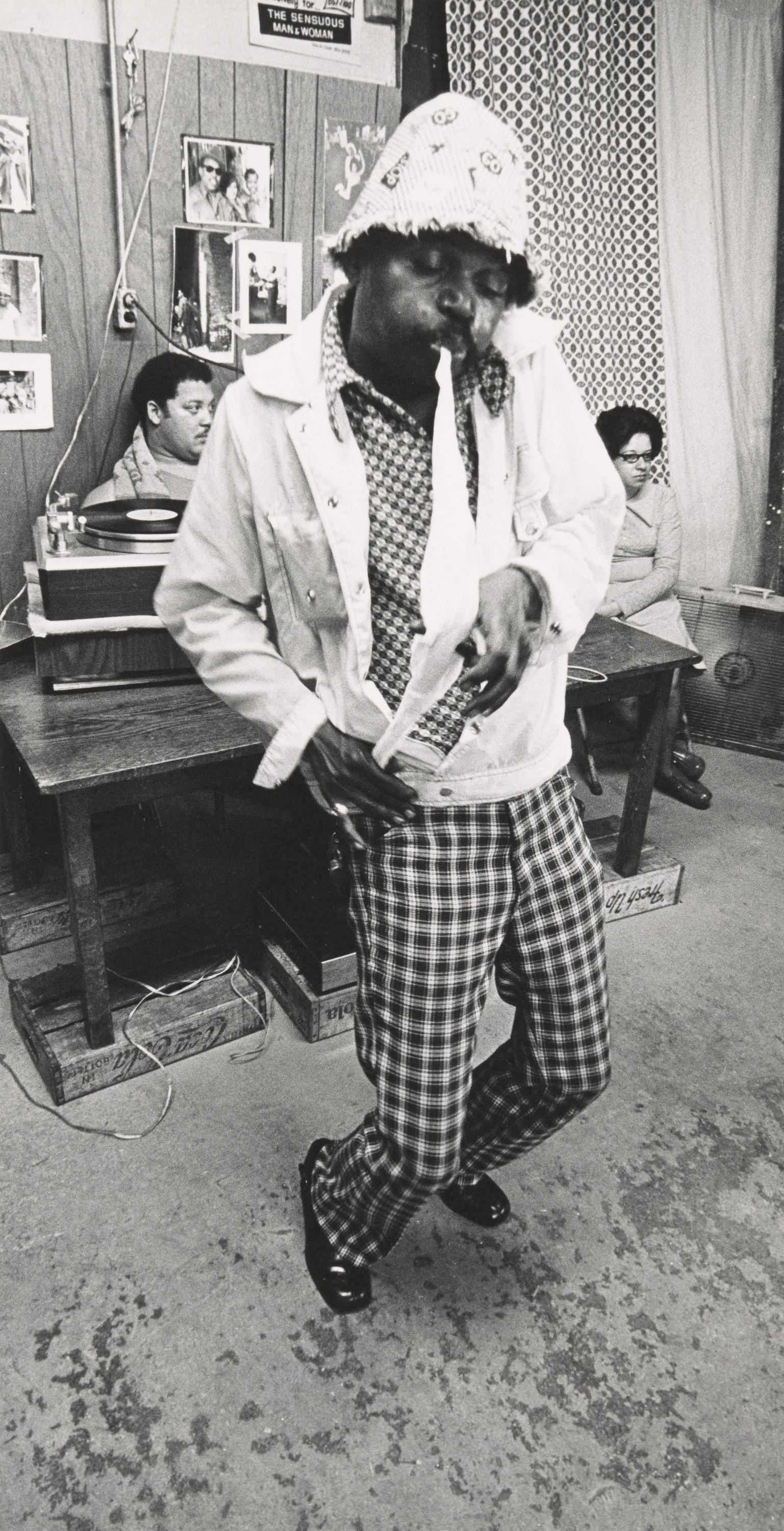 Black-and-white photograph of a man dancing indoors on a scuffed floor. He wears a patterned bucket hat, printed button-down shirt, light-colored jacket, plaid pants, and shiny black shoes. He leans forward, mid-movement, with a focused expression and slightly pursed lips. Behind him, a man and a woman sit at separate wooden desks, and a record player is visible to the left. The wall is decorated with family photographs, posters, and a patterned curtain.