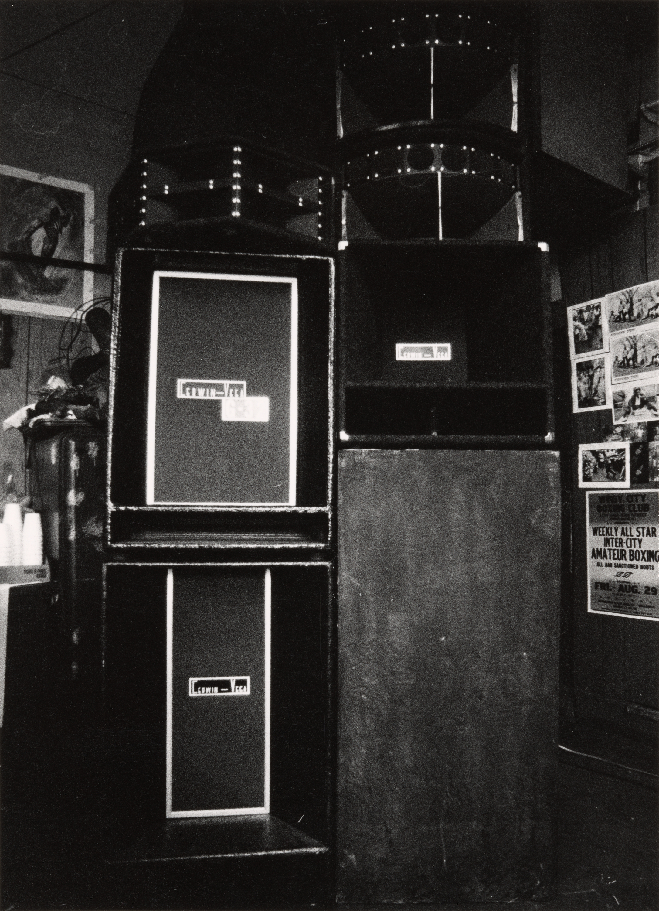 Black-and-white photograph of two tall, stacked speaker cabinets with visible "Cerwin-Vega" logos, positioned against a paneled wall. The speakers dominate the frame and appear to be part of a sound system in a community space. On the right wall, several flyers and photographs are pinned, including a visible poster for a weekly amateur boxing event. On the left side, a fan, a drink dispenser, and a stack of plastic cups are partially visible. The image conveys a setting used for gatherings or performances.