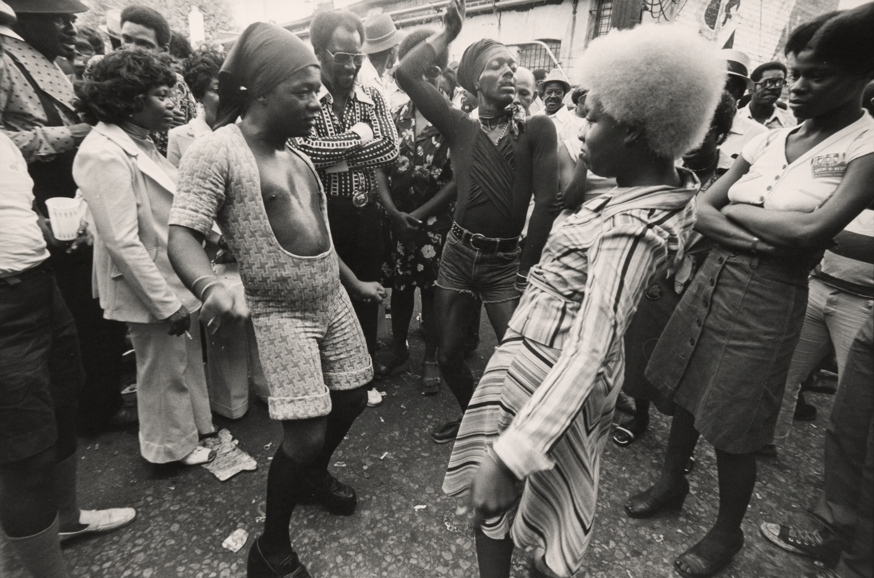 Black-and-white photograph of a lively street scene with people gathered closely together. Three people in the center are dancing with expressive body movements: a person on the left wears a patterned short set and headwrap, a person in the middle wears a black top and shorts with one arm raised, and a person on the right, facing slightly away, wears a striped dress and has a large afro. Surrounding them is a crowd of onlookers, many smiling or watching intently, dressed in 1970s fashion. The street is littered with paper debris, and a building wall with windows is visible in the background.