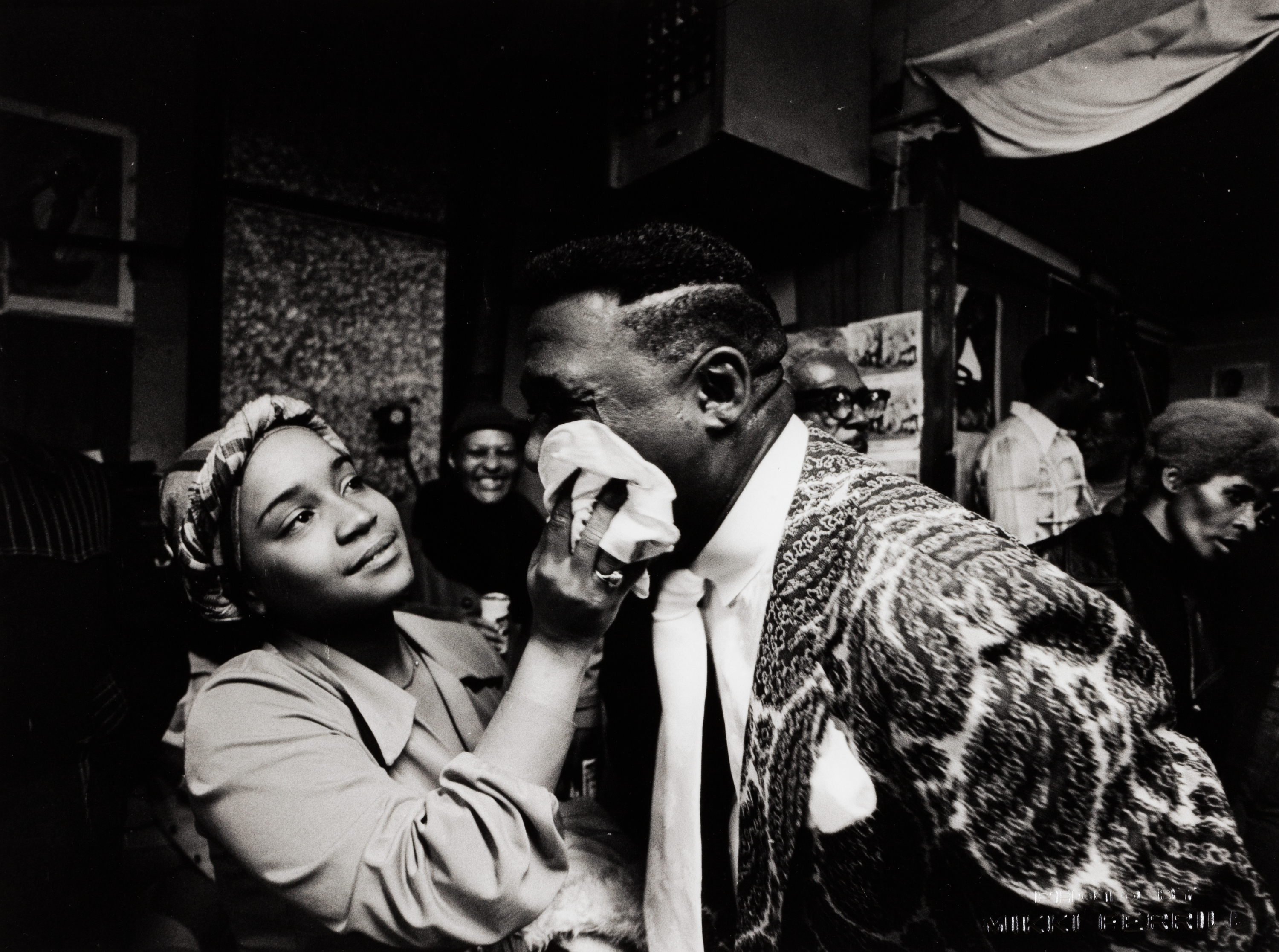 Black-and-white photograph of a woman dabbing a man’s cheek with a cloth as he smiles, wearing a patterned jacket and white tie. The woman, wearing a headscarf and coat, gazes at him with a soft expression. They are surrounded by other people in a warmly lit, crowded interior. Several figures in the background appear to be watching and smiling. Posters or photographs are visible on the wall behind them.