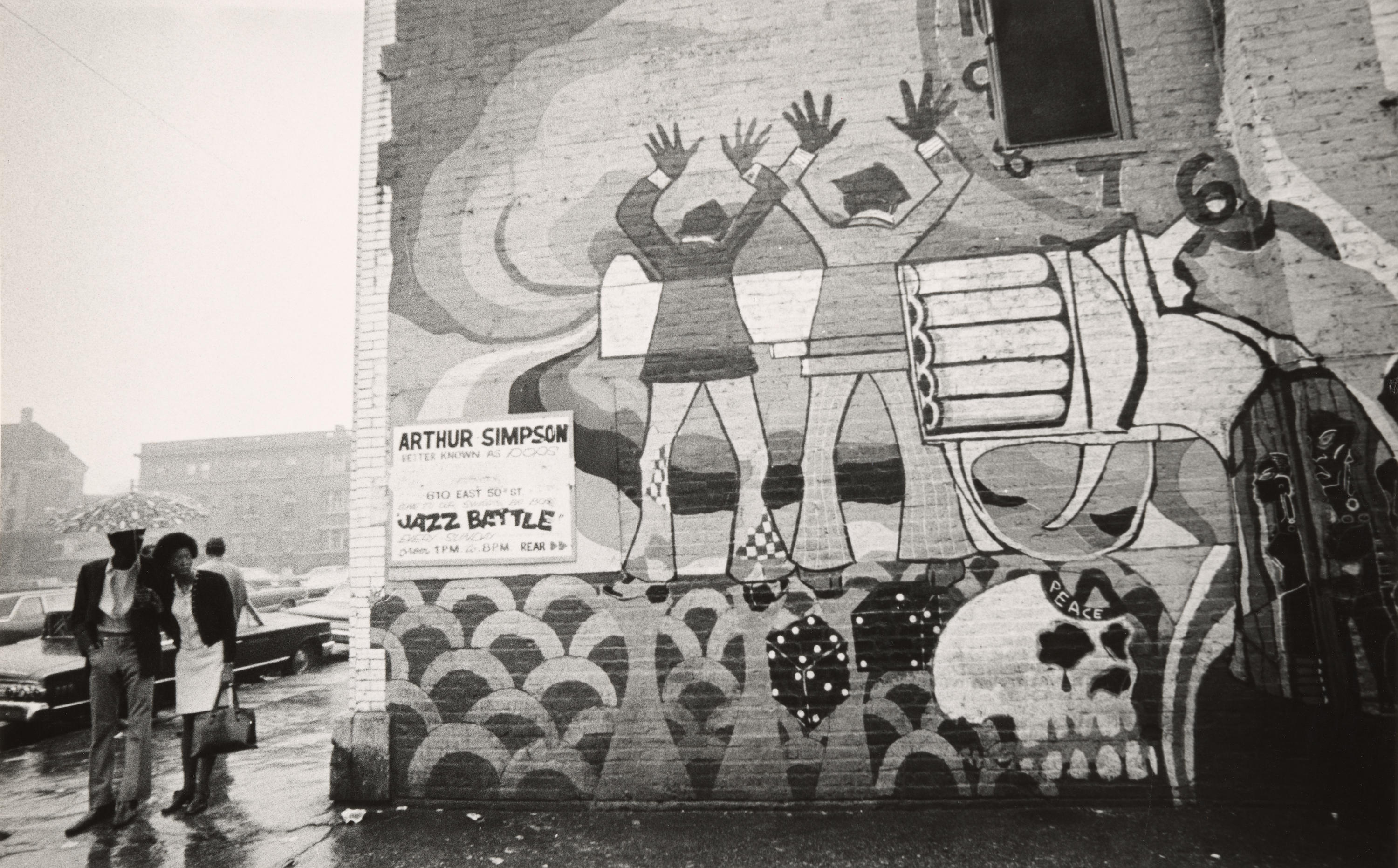 Black-and-white photograph of a painted brick wall featuring a mural with two stylized figures raising their hands in front of an abstract white gun. The figures wear suits and flared pants, and the gun appears to aim toward the right side of the wall, where a skull labeled “PEACE” is incorporated into the design. Beneath the mural is a hand-painted sign reading “Arthur Simpson, better known as Pops, Jazz Battle.” On the sidewalk, a man and woman stand under an umbrella in rainy weather, looking toward the wall. Parked cars and blurred buildings appear in the background.