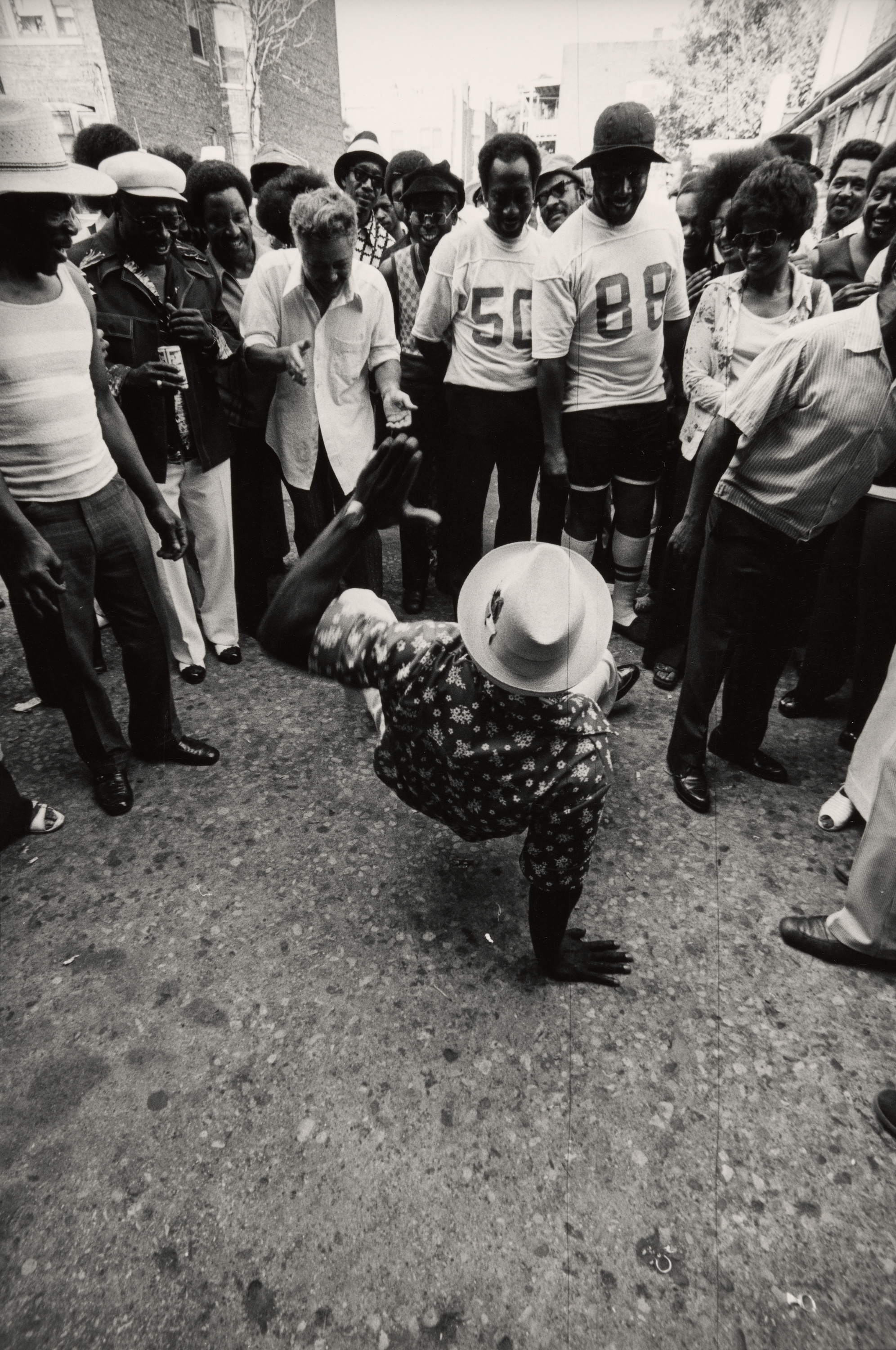 Black-and-white photograph of a man breakdancing on one hand in the center of a circle of onlookers in an outdoor setting. The man wears a patterned shirt and a wide-brimmed hat, with his legs in motion and head low to the ground. Around him, a crowd of people smiles, laughs, and gestures with excitement. Some wear casual clothing, while others are in uniforms or hats. The street is paved with worn concrete, and buildings and trees are visible in the background. The atmosphere is festive and energetic.