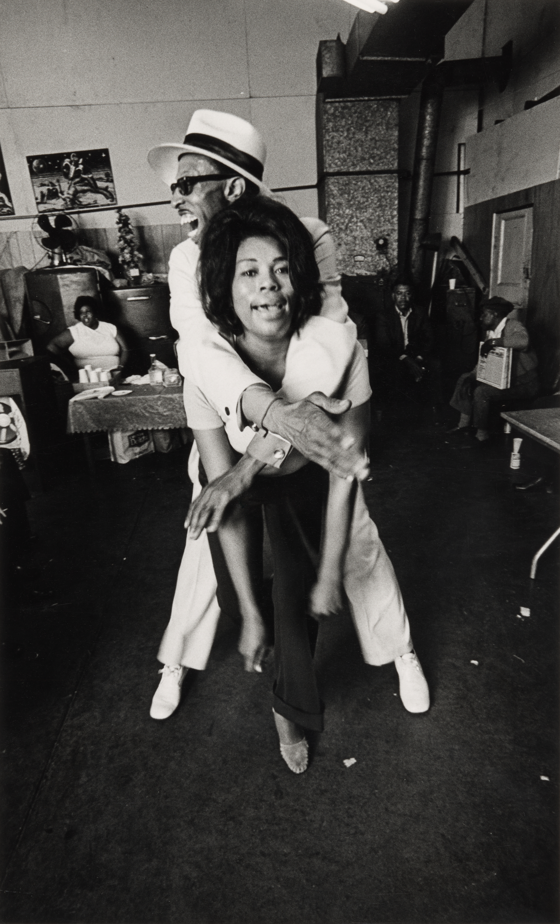 Black-and-white photograph of a joyful moment inside a community space. In the center, a man in a white suit and hat wraps his arms around a laughing woman from behind, both of them caught mid-motion as they dance. The woman leans forward with a wide smile, and the man grins over her shoulder. Behind them, people sit and watch, including a woman at a table on the left and two seated men on the right. Posters and decorations hang on the wood-paneled walls, adding to the informal, celebratory atmosphere.