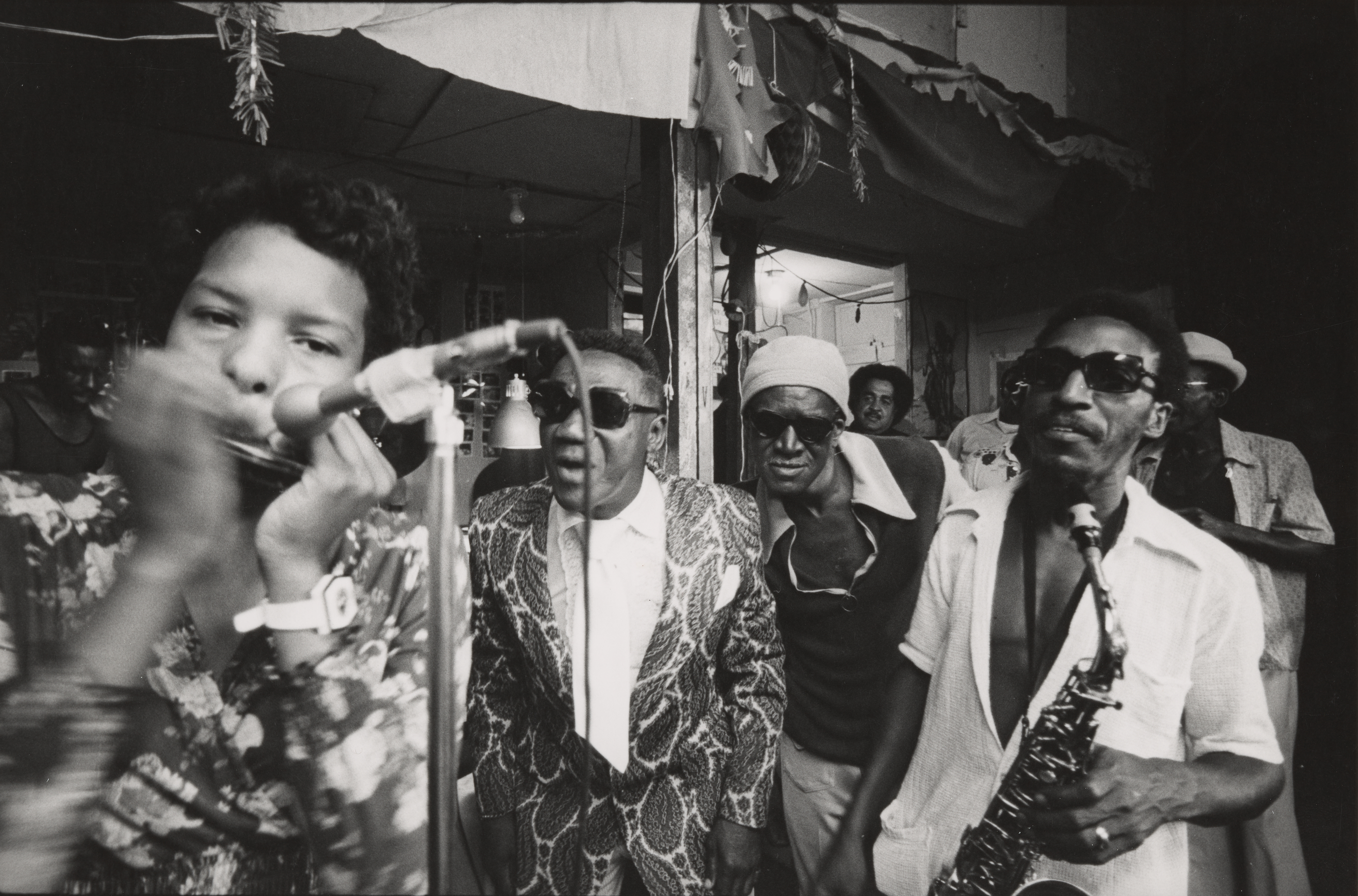 Black-and-white photograph of a musical performance in an indoor setting. In the foreground at left, a woman plays harmonica into a microphone, her face close to the lens. Behind her, a man in a patterned suit and dark sunglasses sings into a second microphone. Next to him, two more men, one wearing a headscarf and the other a white short-sleeved shirt and sunglasses, stand closely together, the latter holding a saxophone. Other people fill the background in a lively, crowded space decorated with hanging fabric and light fixtures. The atmosphere is energetic and expressive.