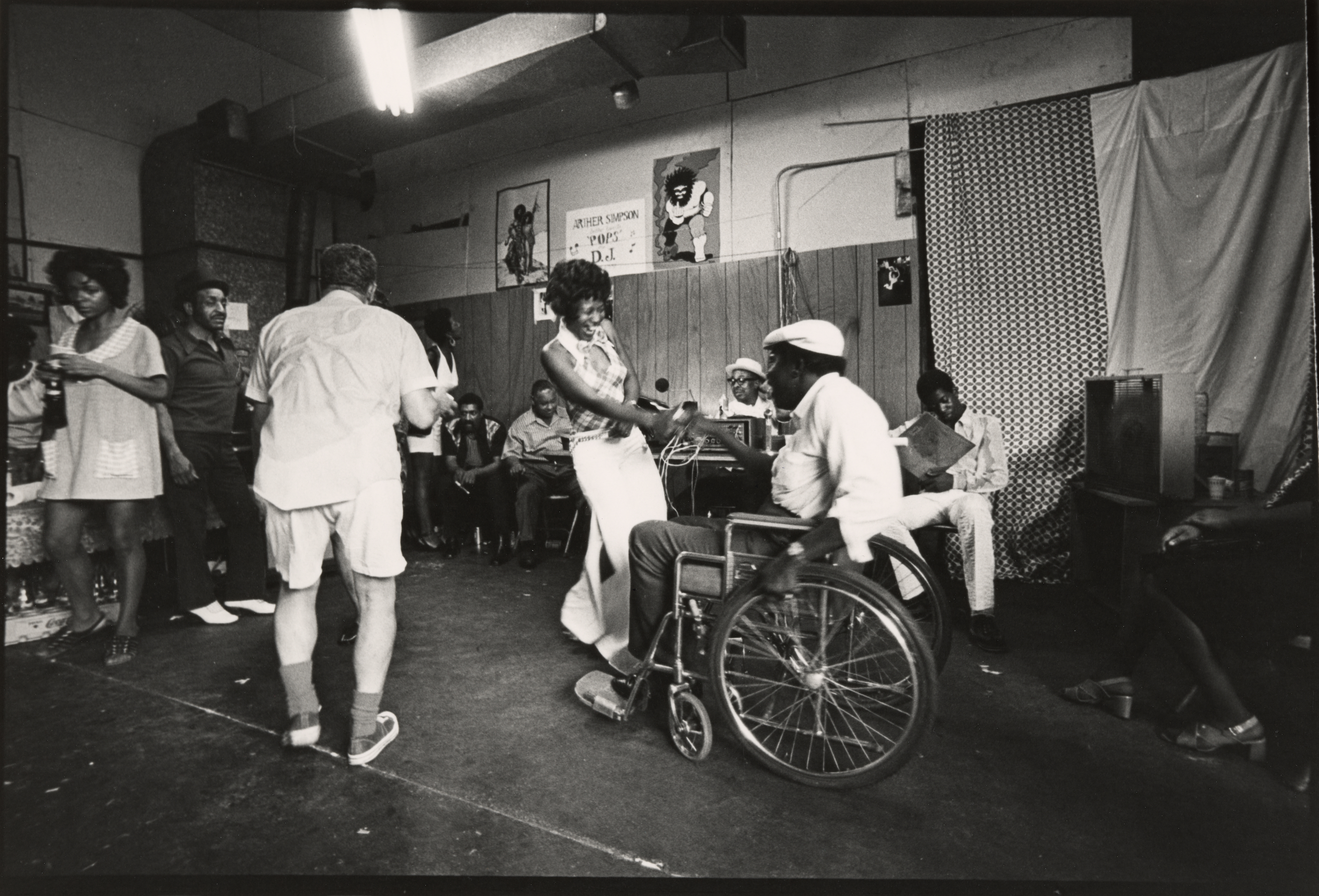 Black-and-white photograph of a lively social gathering inside a wood-paneled room. At the center, a woman in a halter top and long skirt dances with a man seated in a wheelchair; they hold hands and smile. Other people stand or sit around the edges of the room, watching, talking, or holding drinks. Posters and patterned fabric decorate the walls, and a man in the background sits behind a turntable setup. A handwritten sign reads "Arthur Simpson 'Pops' D.J." above the dancers.