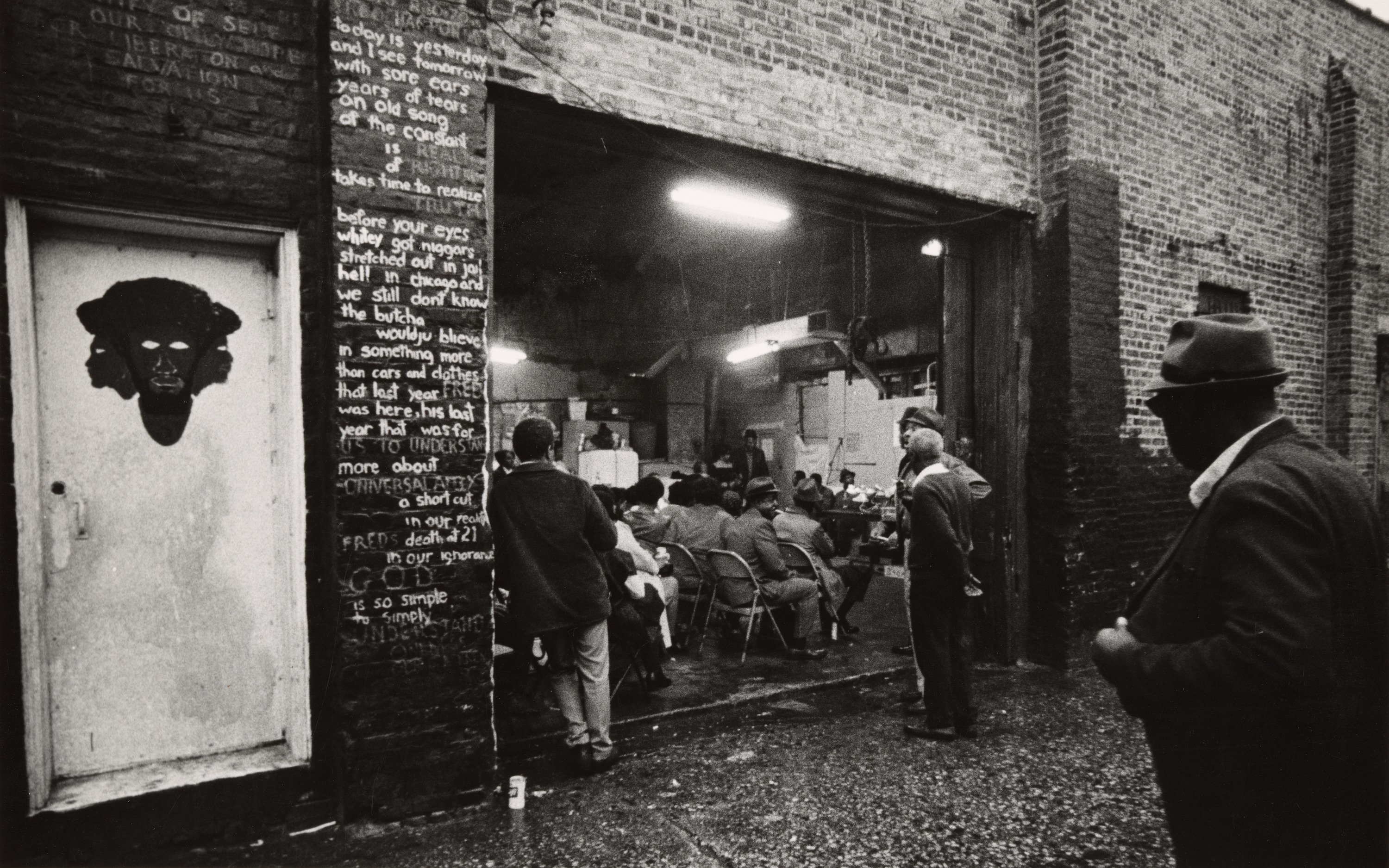 Black-and-white photograph showing an open brick garage filled with seated people, appearing to listen to a speaker near the back. The garage door is raised, and fluorescent lights illuminate the interior. Outside, two men stand in the foreground on a wet pavement, one in shadow and another with his back to the camera. To the left, a painted door features a stylized Black face, and adjacent bricks are covered with chalk-written poetry or text referencing incarceration and a person named Fred. The scene suggests a community gathering or meeting taking place in an informal setting.